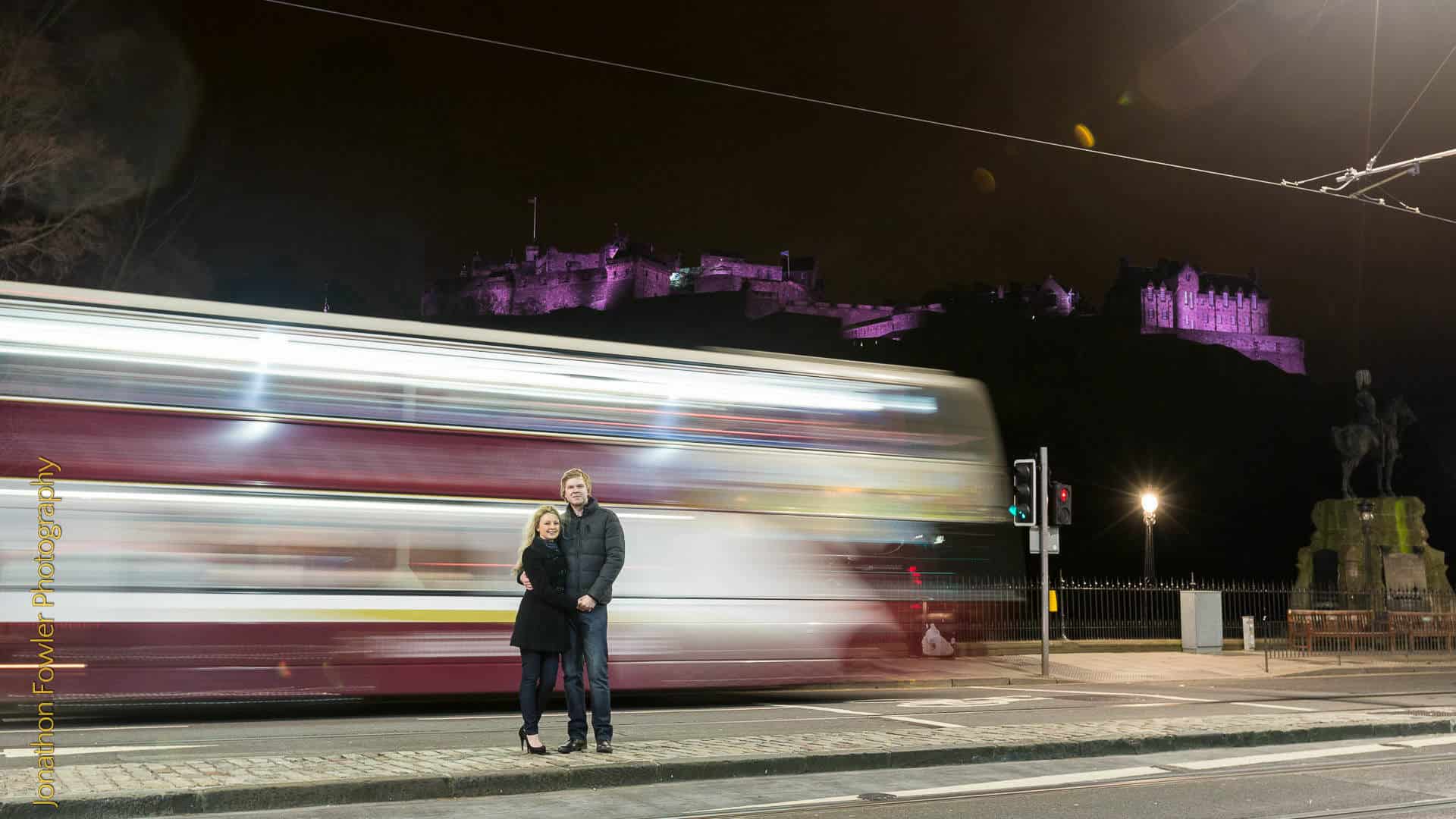 Edinburgh city centre night-time pre-wedding photoshoot Andrew and Nicola: A couple stands on a sidewalk at night with a blurred double-decker bus passing by. In the background, a castle is illuminated in purple light.