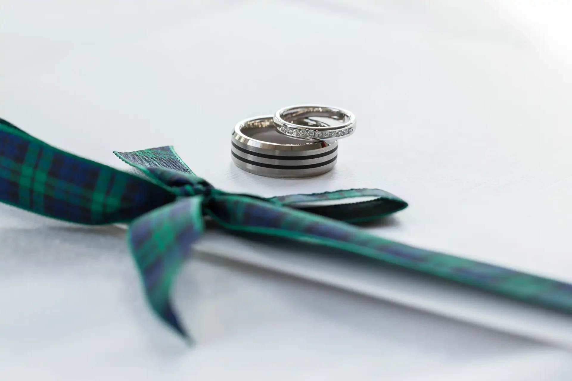 Two wedding rings stacked beside a green and blue tartan bow tie on a white fabric background.