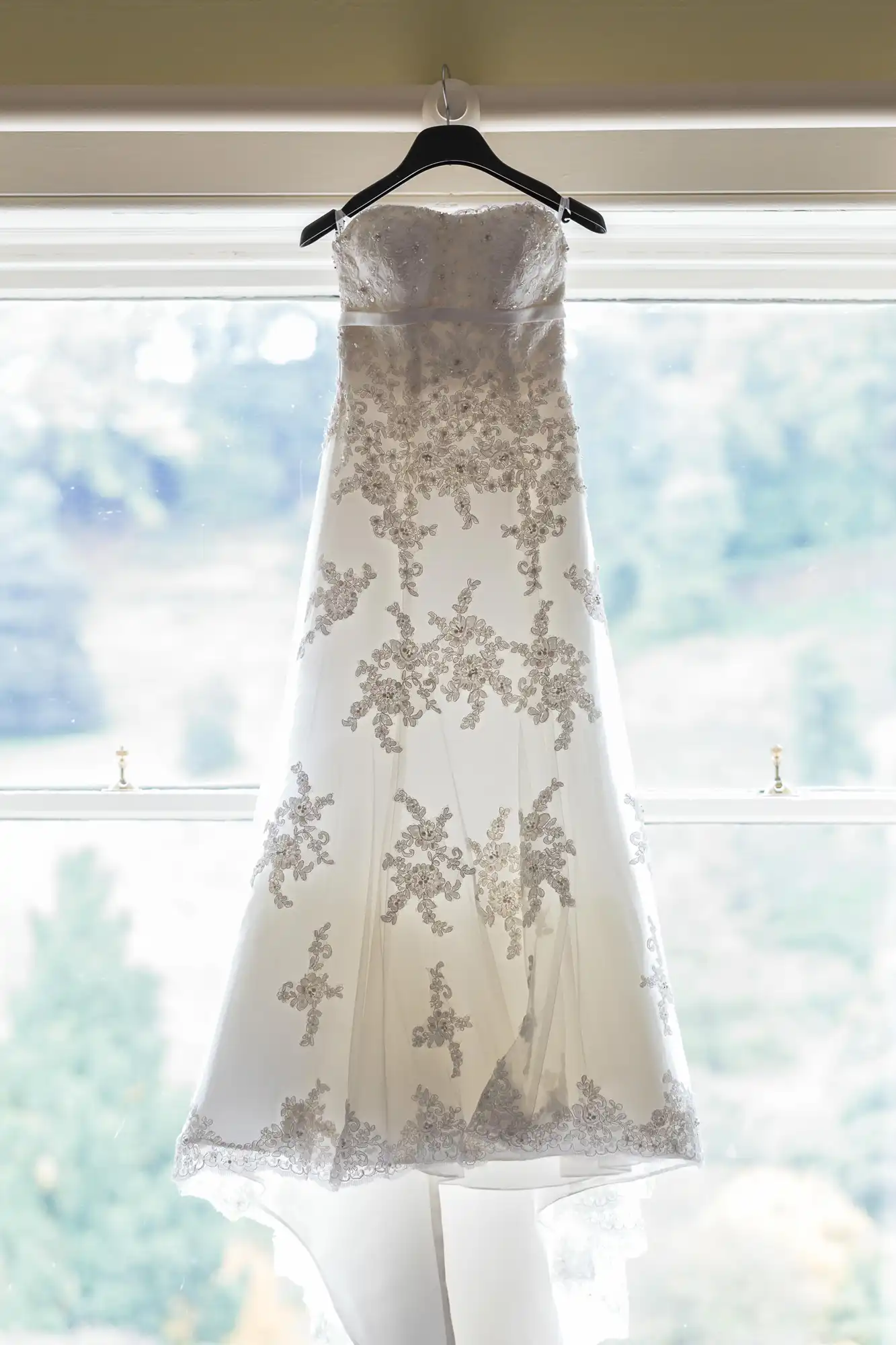 A white bridal gown with lace details hanging in front of a window, showcasing a scenic outdoor backdrop.