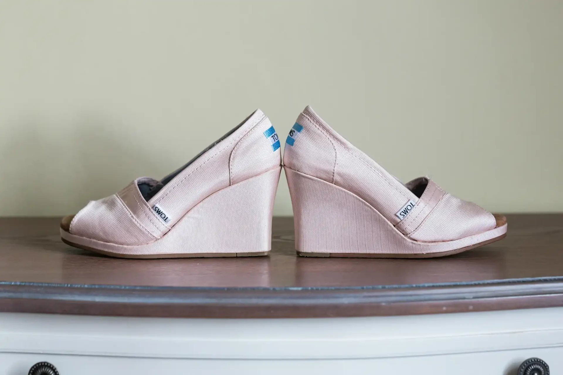A pair of beige wedge heel shoes with "toms" labels, displayed on a wooden surface against a green wall.