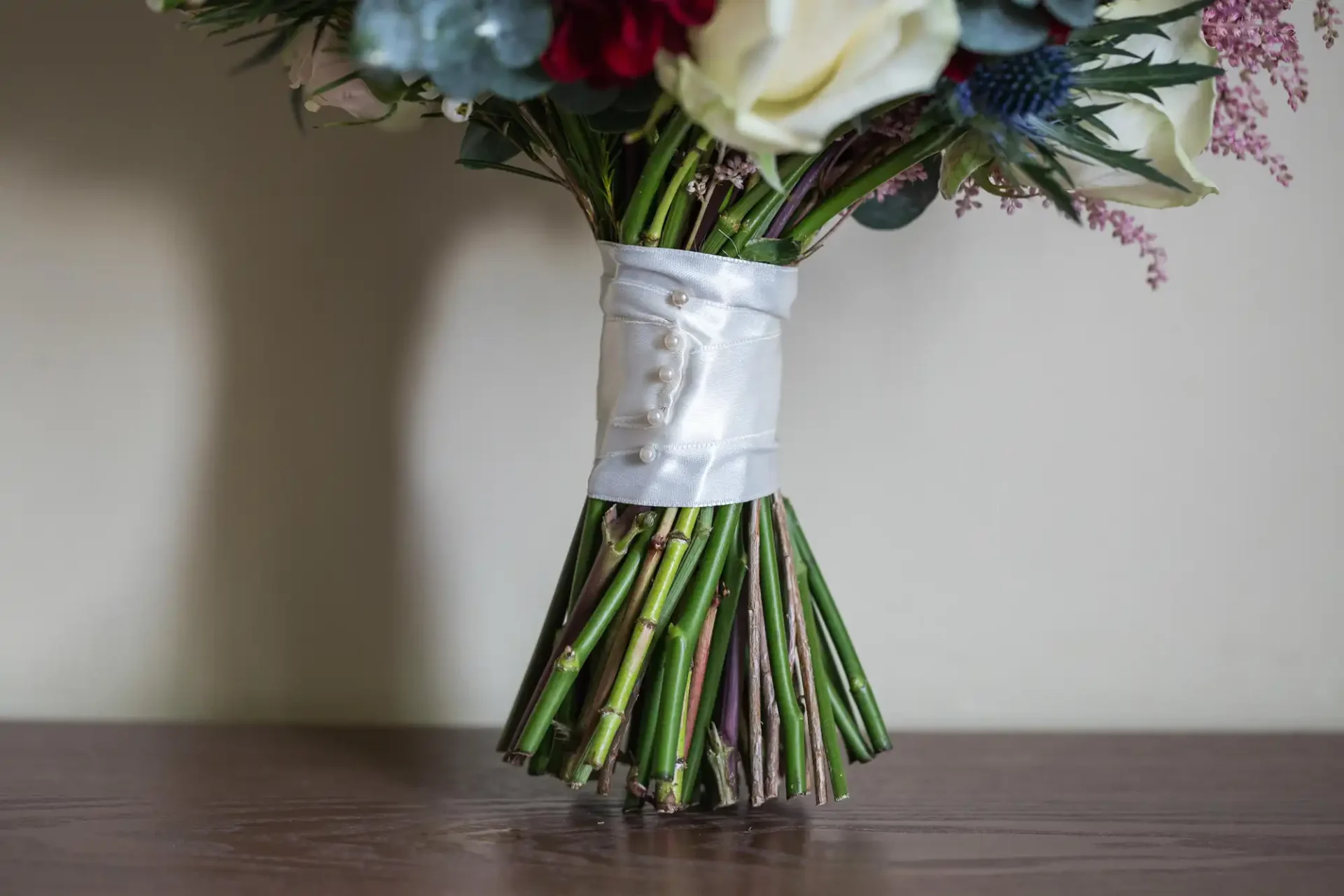 A bridal bouquet with a white satin ribbon handle decorated with pearl buttons, placed on a wooden surface.