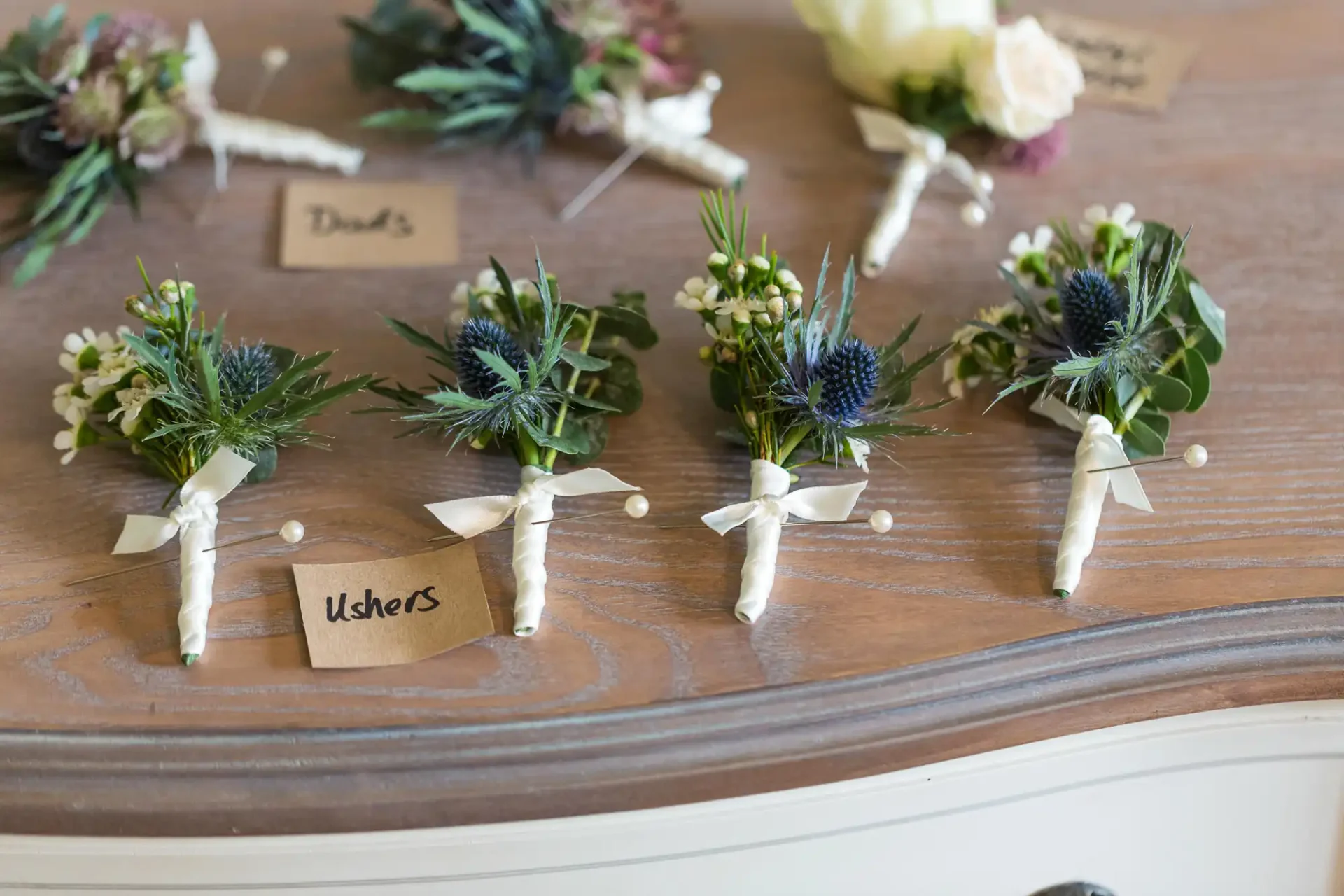 Boutonnieres with tags labeled "dads" and "ushers" laid out on a wooden surface.