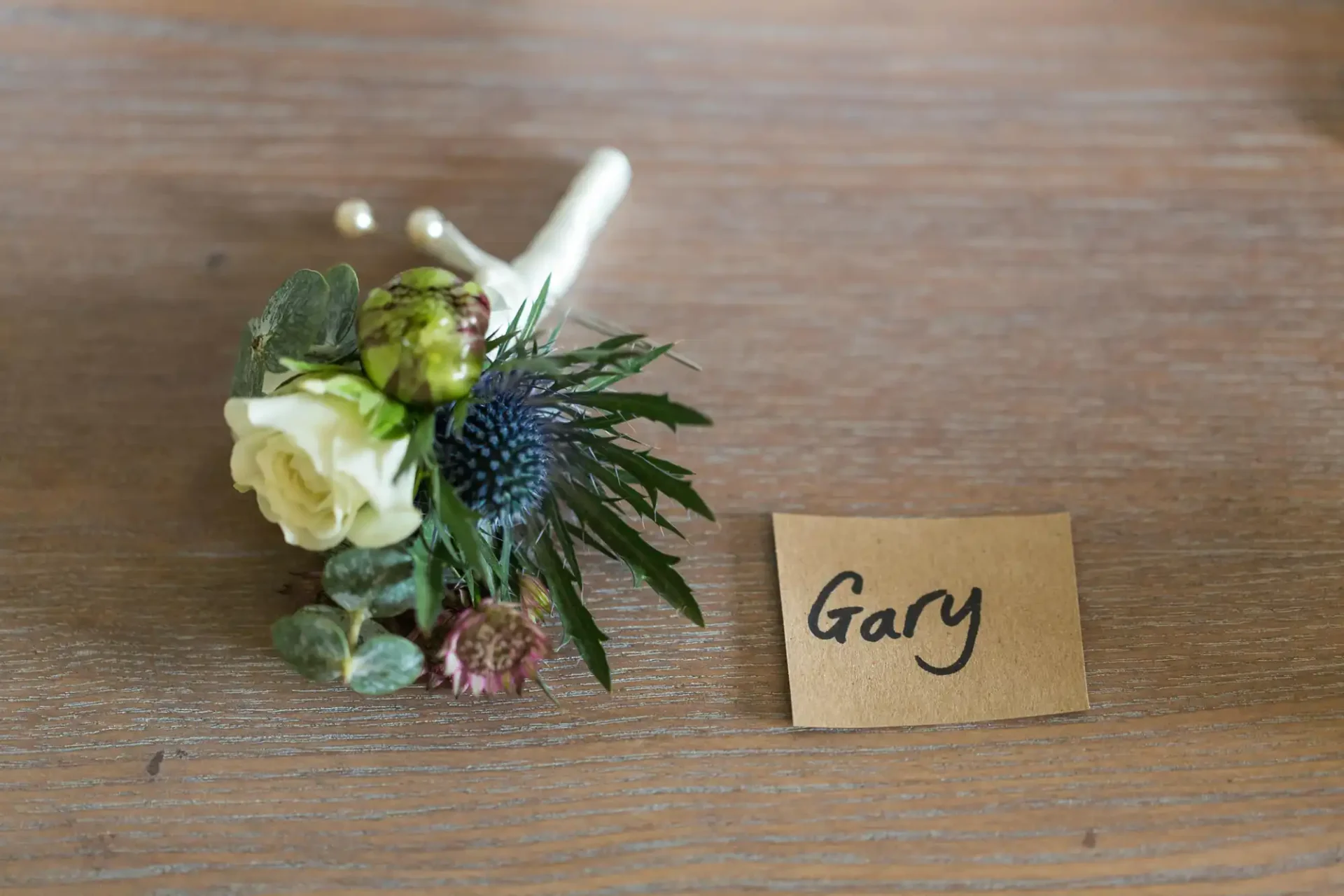 A small floral arrangement with a white rose beside a place card labeled "gary" on a wooden table.