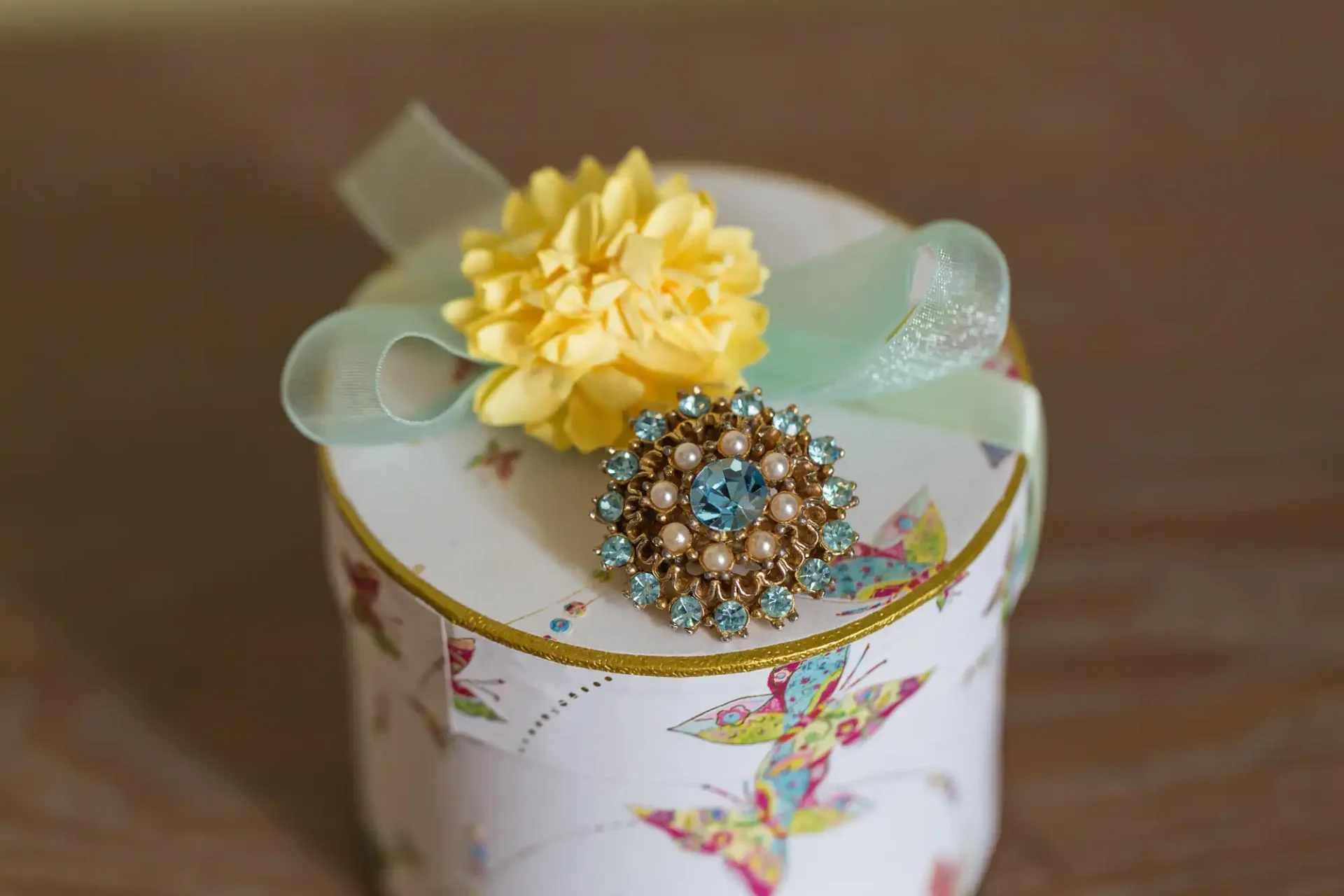A decorative round box with floral prints, adorned with a light blue and gold brooch and a yellow flower on top, tied with a pale green ribbon.