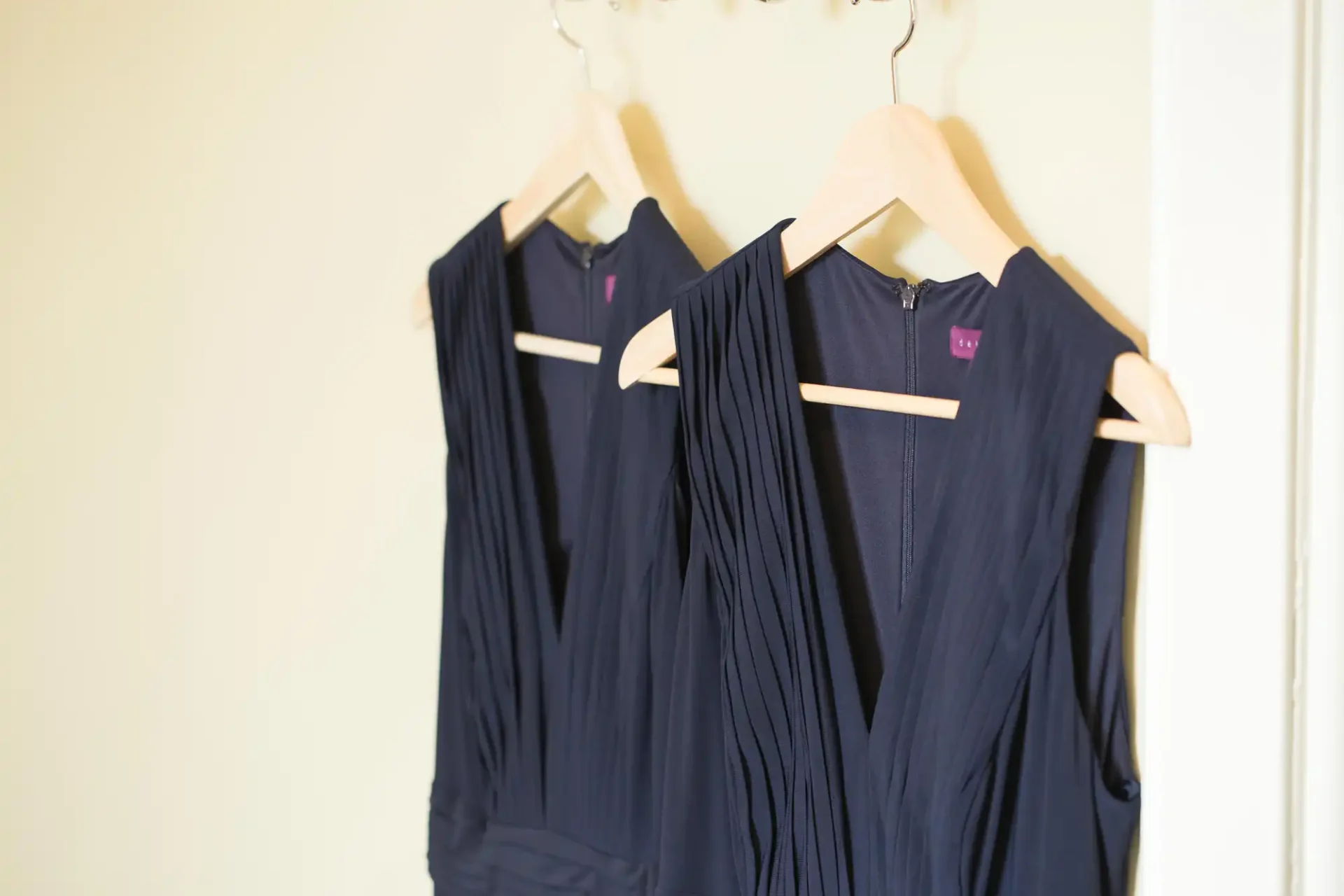Two navy blue dresses hanging on wooden hangers against a pale yellow background.