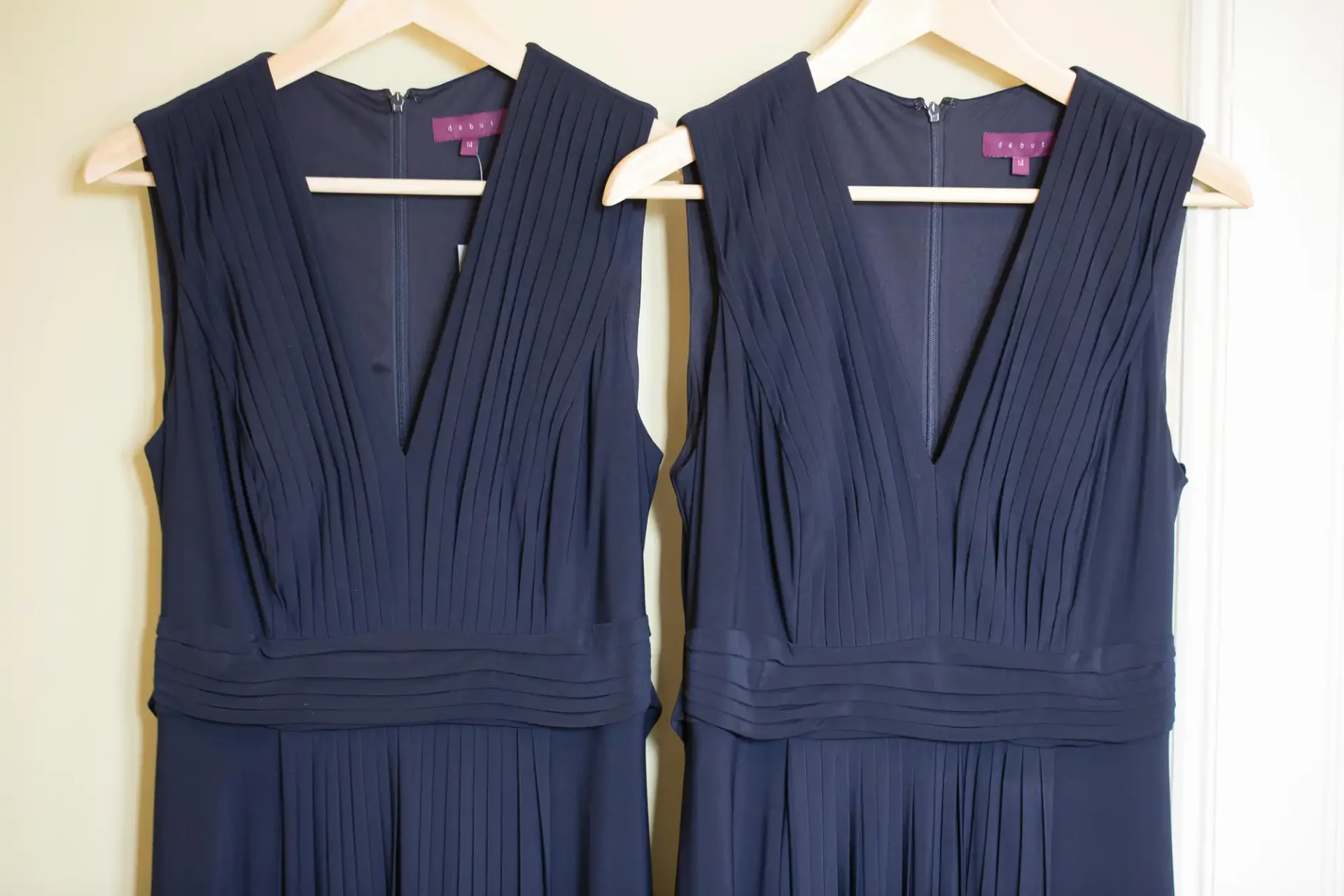 Two navy blue dresses with pleated designs hanging on white hangers against a cream-colored door.