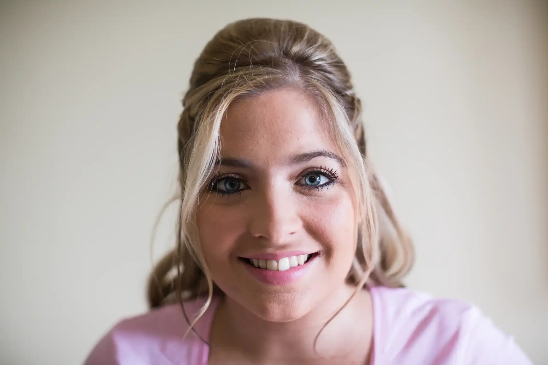 Close-up portrait of a smiling woman with blonde hair styled in an updo, wearing a pink shirt, against a light background.