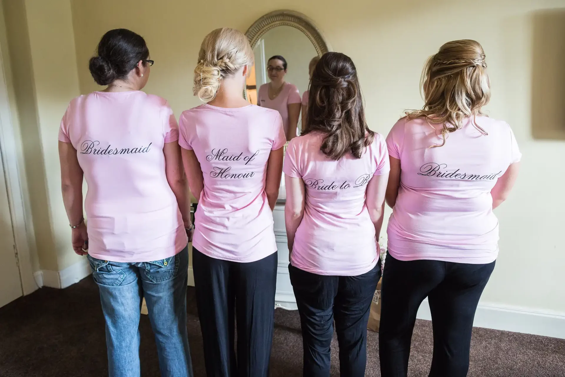 Four women stand facing away, wearing pink t-shirts labeled "bridesmaid" and "maid of honour," looking at a mirror reflection.