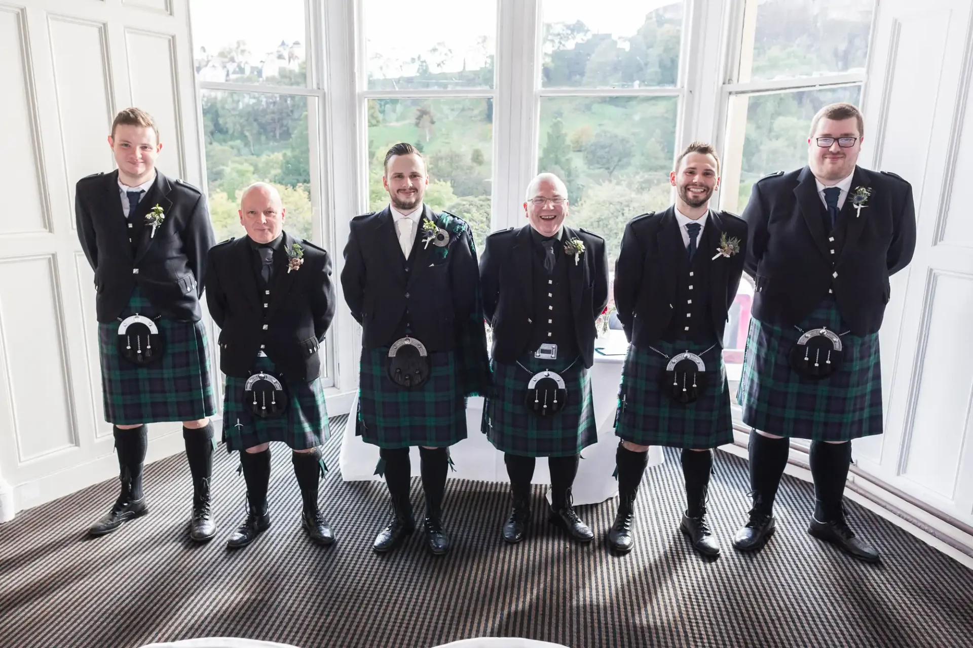 Six men in scottish kilts and formal jackets smiling indoors in front of large windows with a view of greenery.
