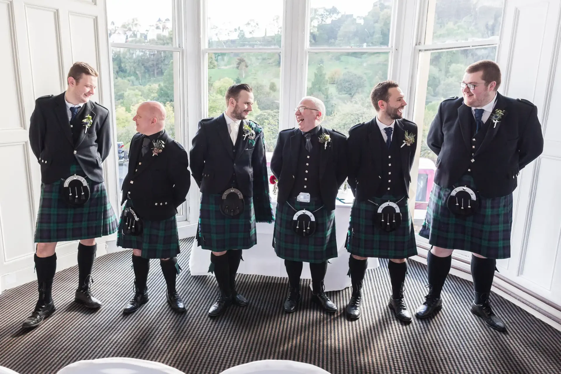 Group of six men in traditional scottish kilts and formal jackets, laughing together by large windows in a bright room.