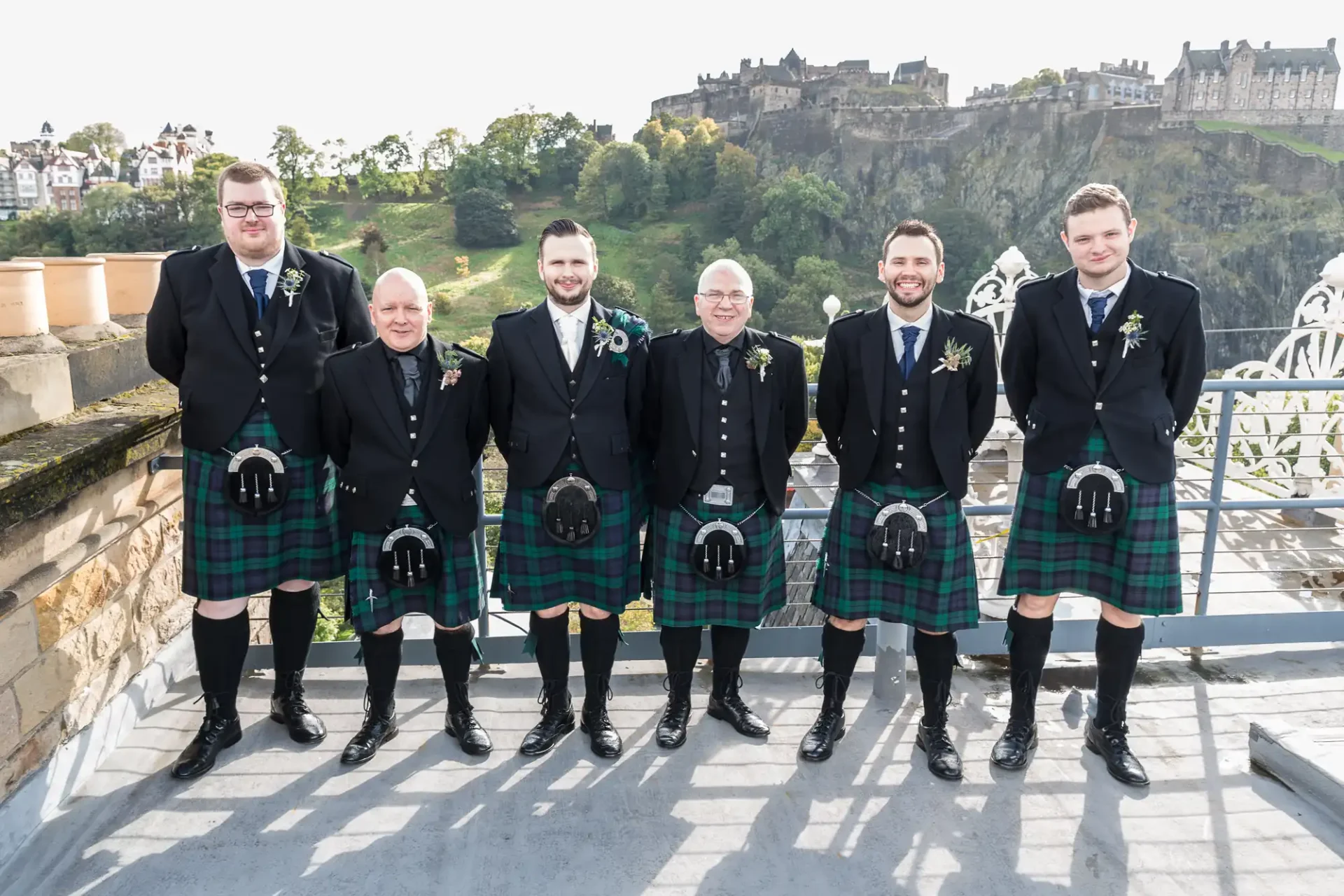 Seven men in traditional scottish kilts standing on a terrace with edinburgh castle in the background.