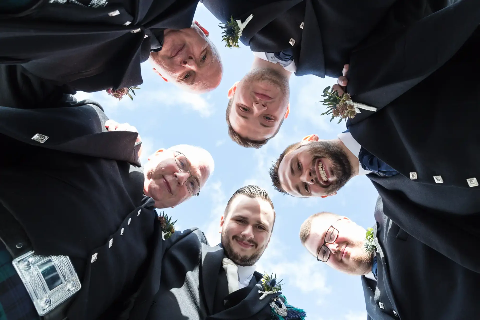 Six men in formal attire with boutonnieres seen from a low-angle, forming a circle with their heads leaning in, against a blue sky with clouds.