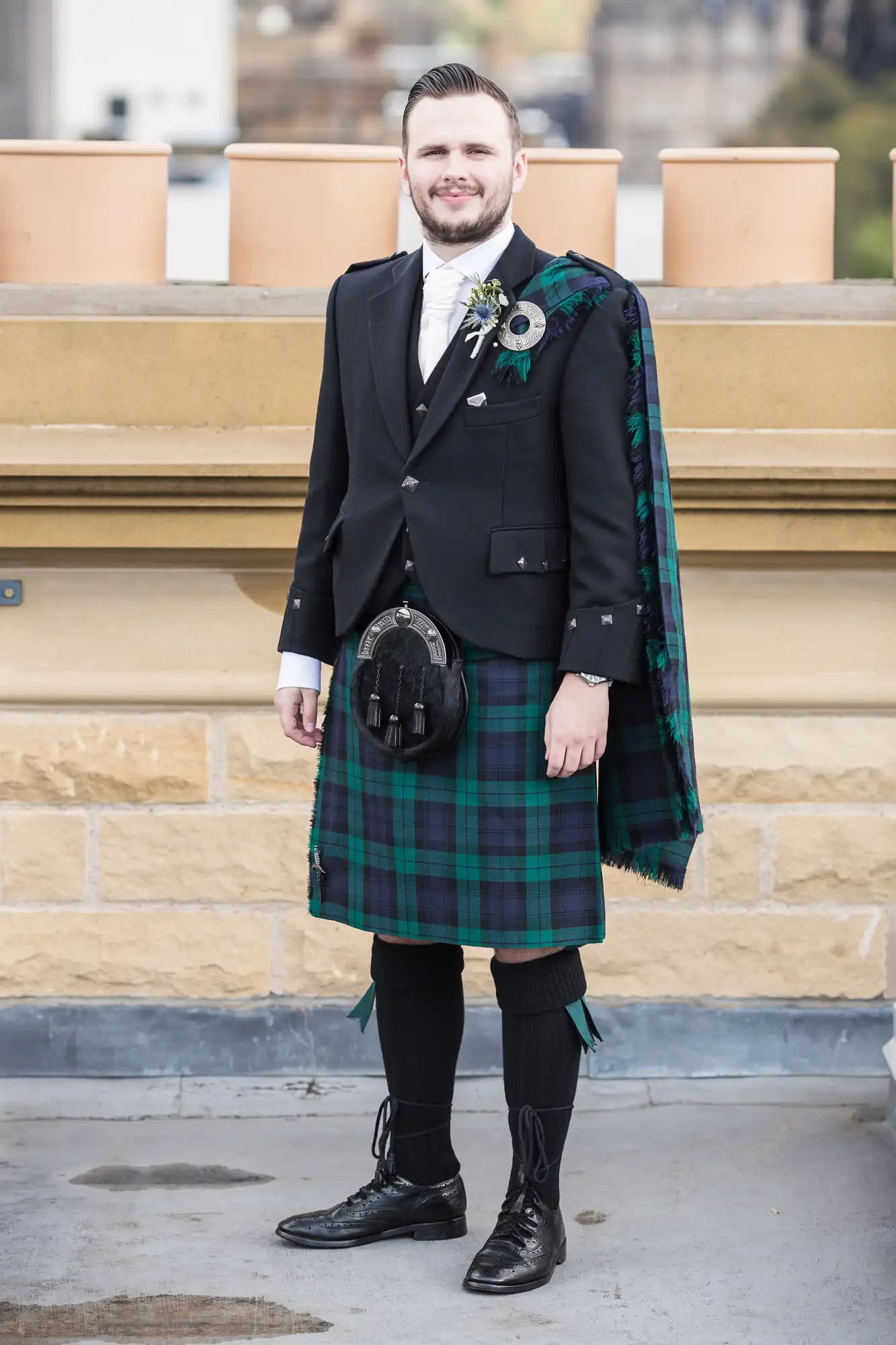 Man in traditional scottish attire, including a tartan kilt and sporran, smiling outdoors.