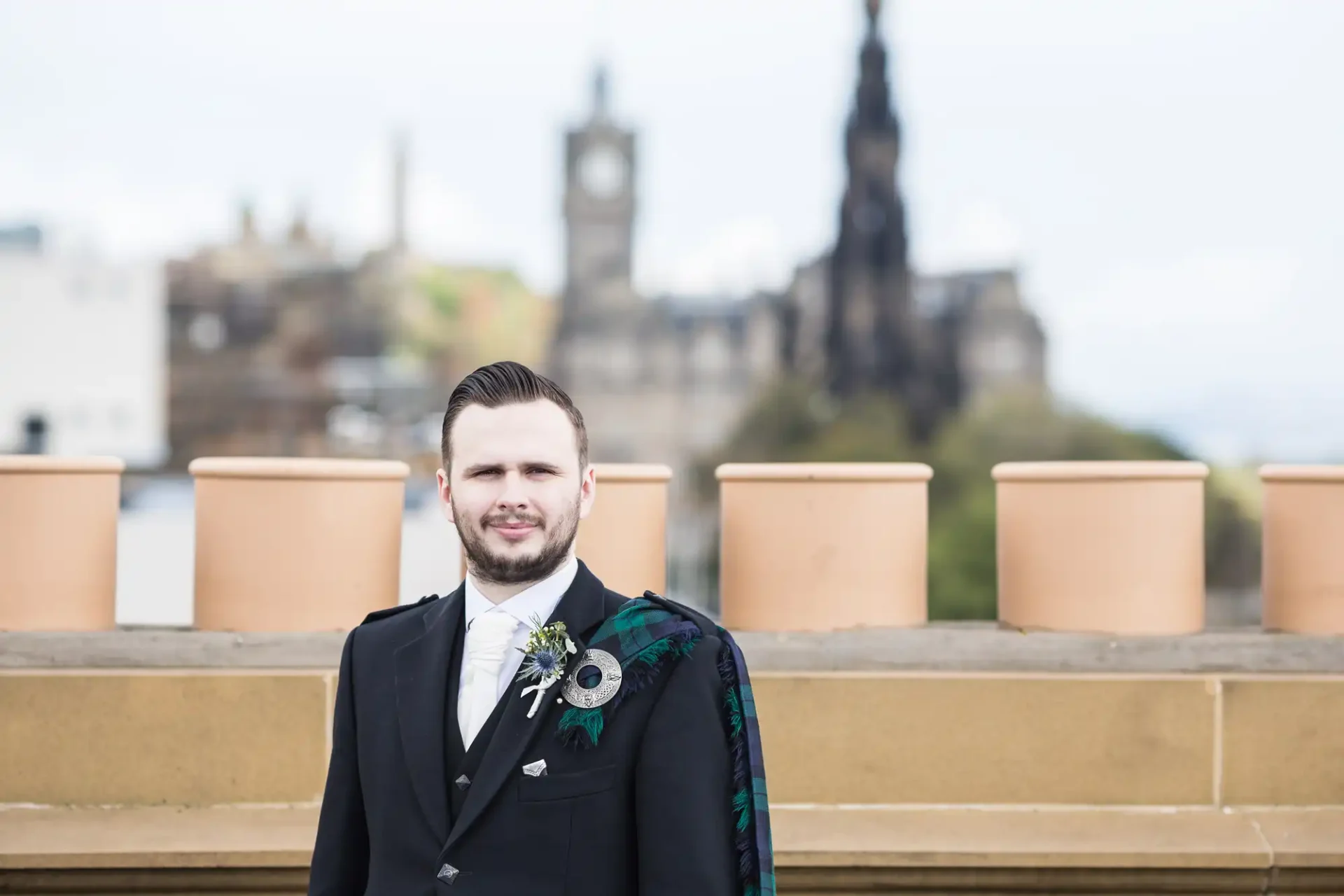 A man in a formal black suit and kilt standing in front of a historical cityscape with a castle in the background.