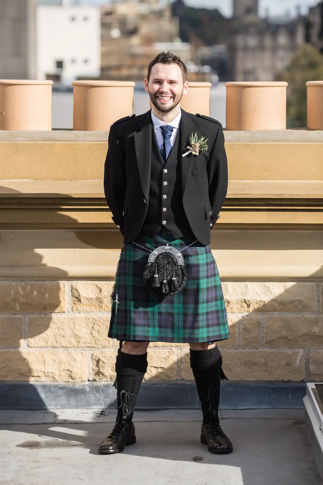 Man in traditional scottish attire including a kilt, sporran, and jacket, smiling on a balcony with buildings in the background.