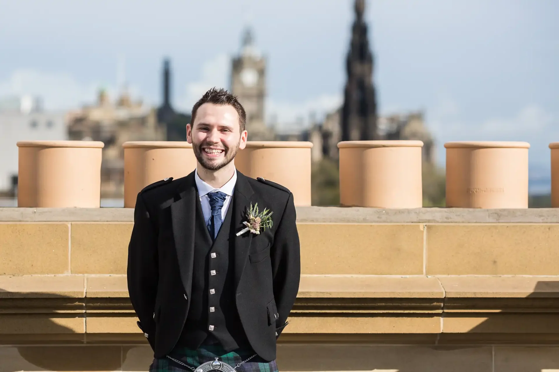 A man in a kilt and black jacket smiling, standing on a balcony with large pots and a cityscape with monuments in the background.