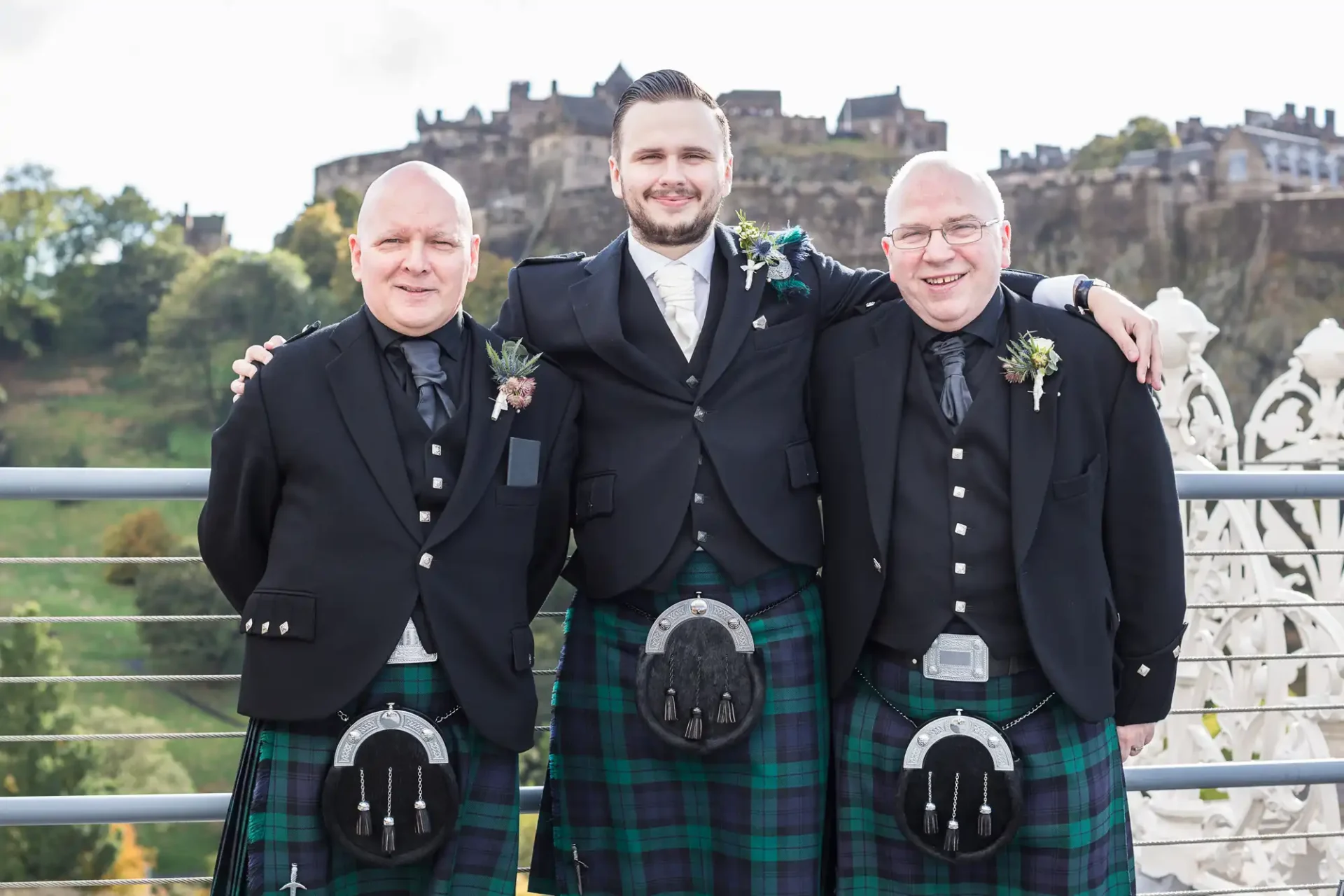 Three men in traditional scottish kilts posing together with edinburgh castle in the background.
