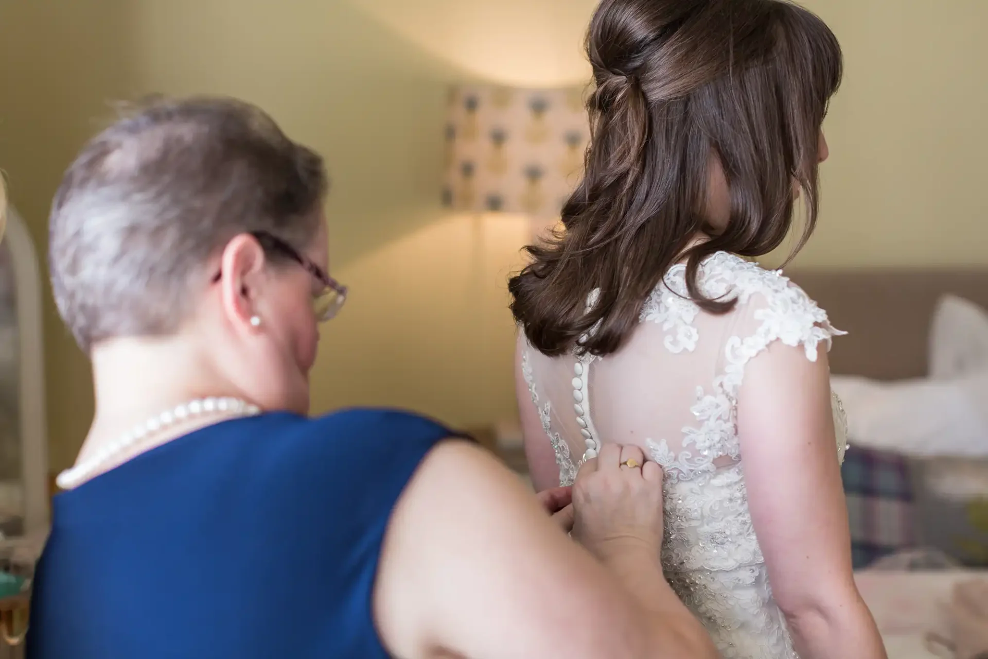 A woman in a blue dress is helping a bride, seen from behind, with the final buttoning of her detailed lace wedding gown in a softly lit room.