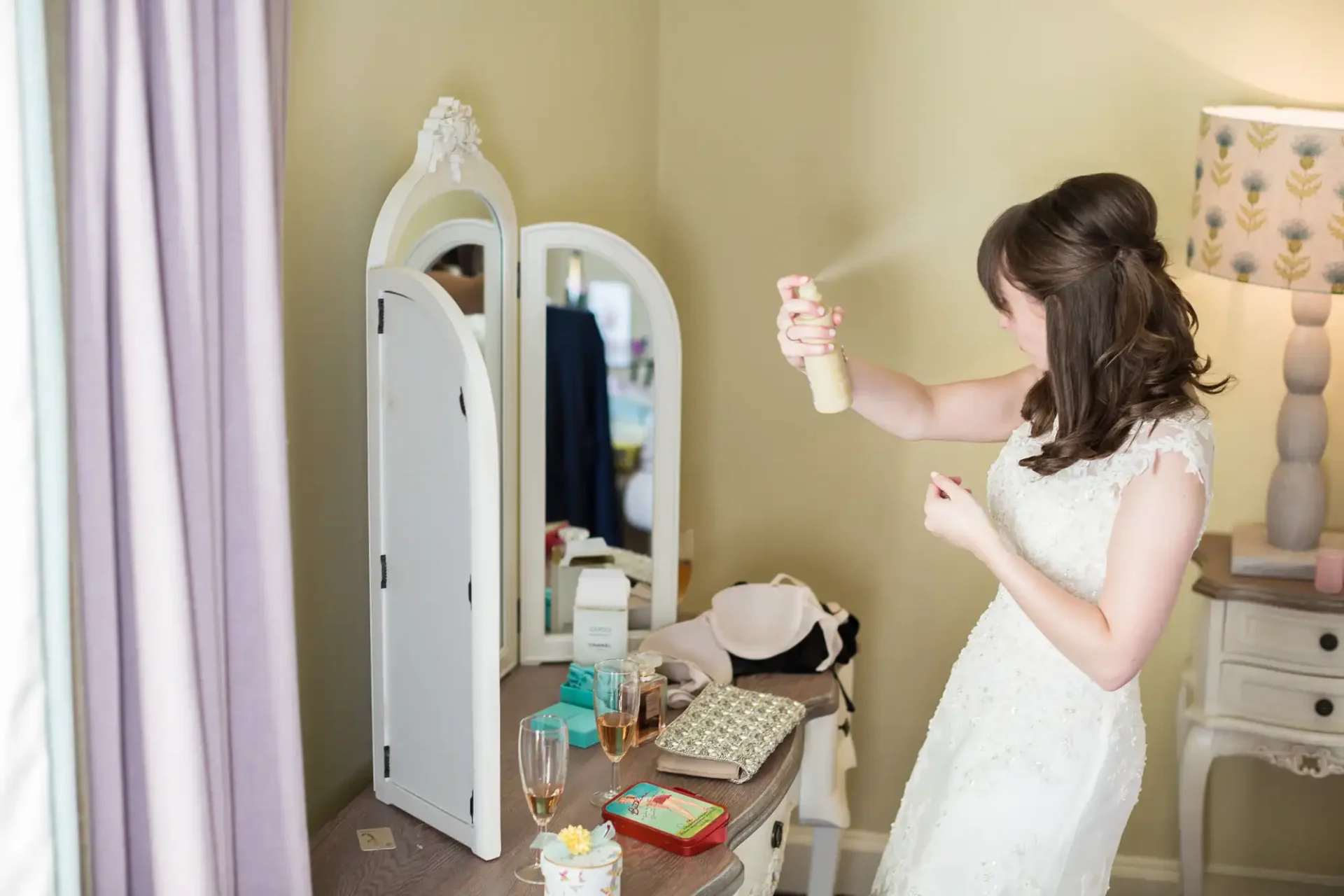 A bride in a white dress applying perfume in front of a mirror in a softly lit room adorned with elegant furniture.