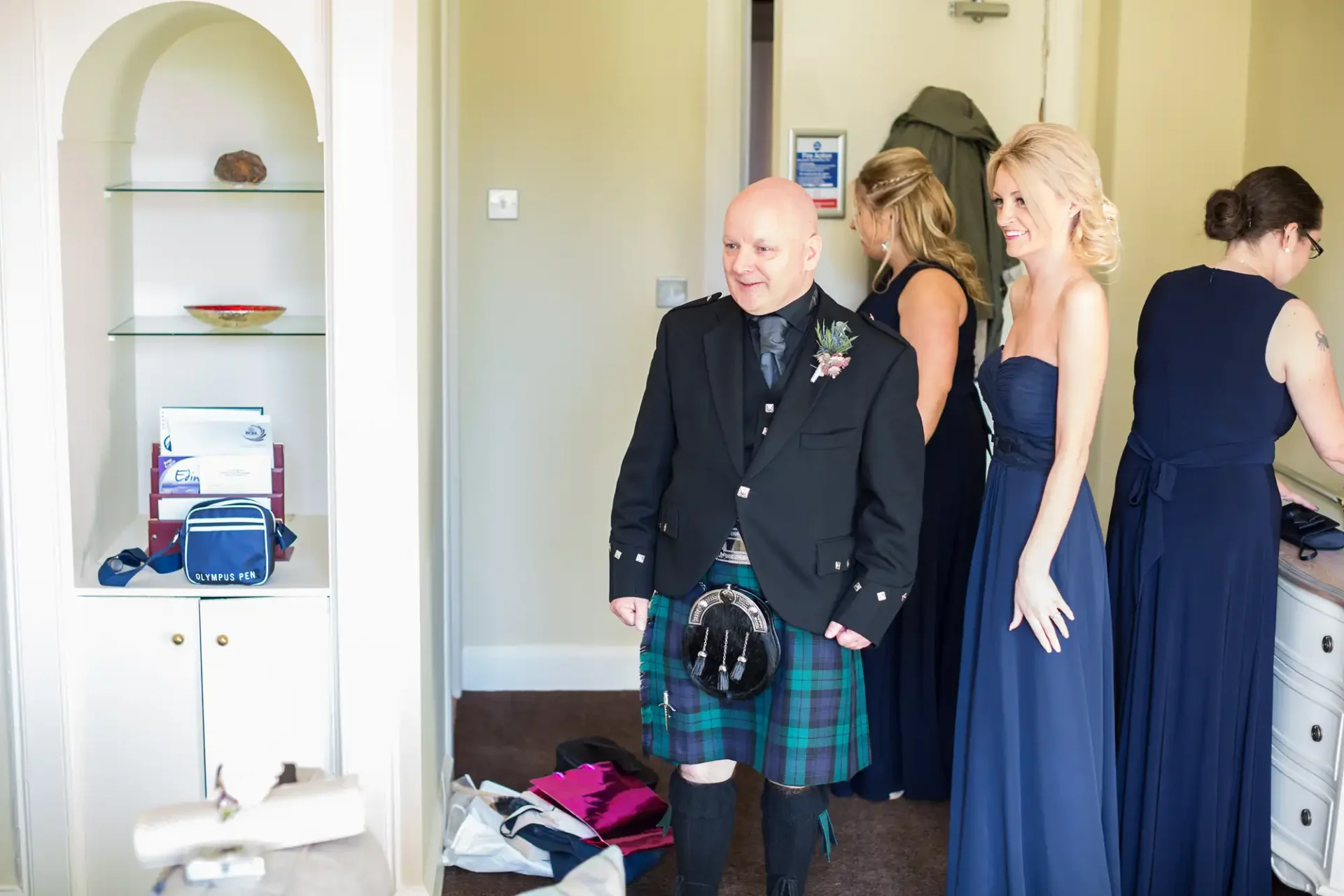 A bald man in a kilt and women in formal dresses in a room, clutter visible on the floor and furniture.