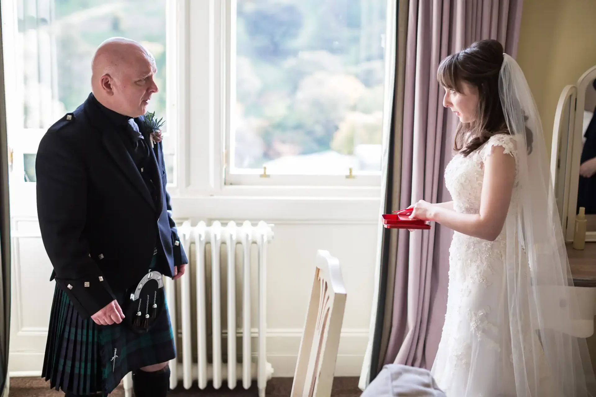 Bride in a white dress presenting a gift to a man in a kilt and formal jacket indoors, with a picturesque view from the window.