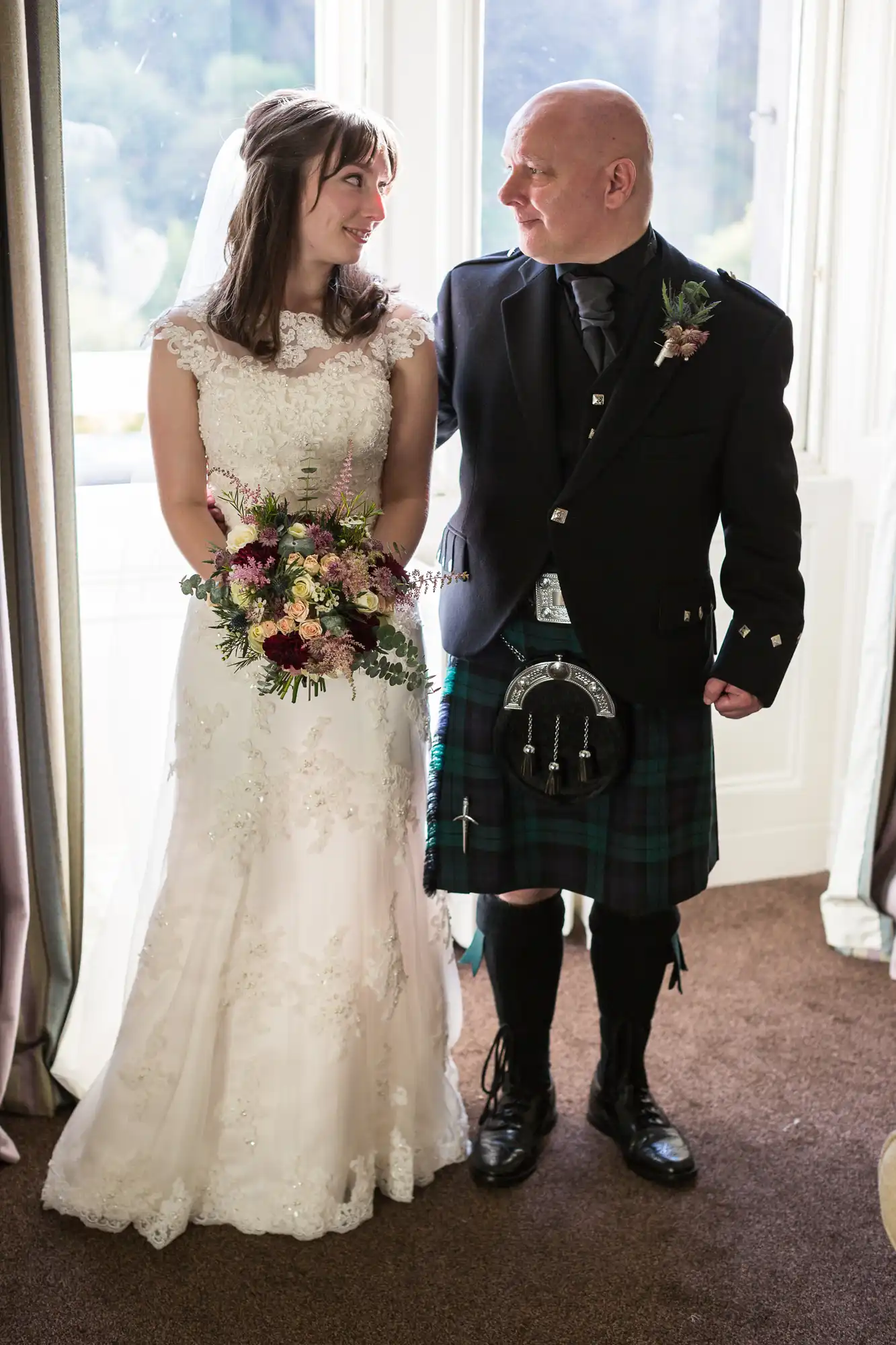 A bride in a lace wedding gown and a groom in a traditional scottish kilt and sporran sharing a joyful moment indoors by a window.