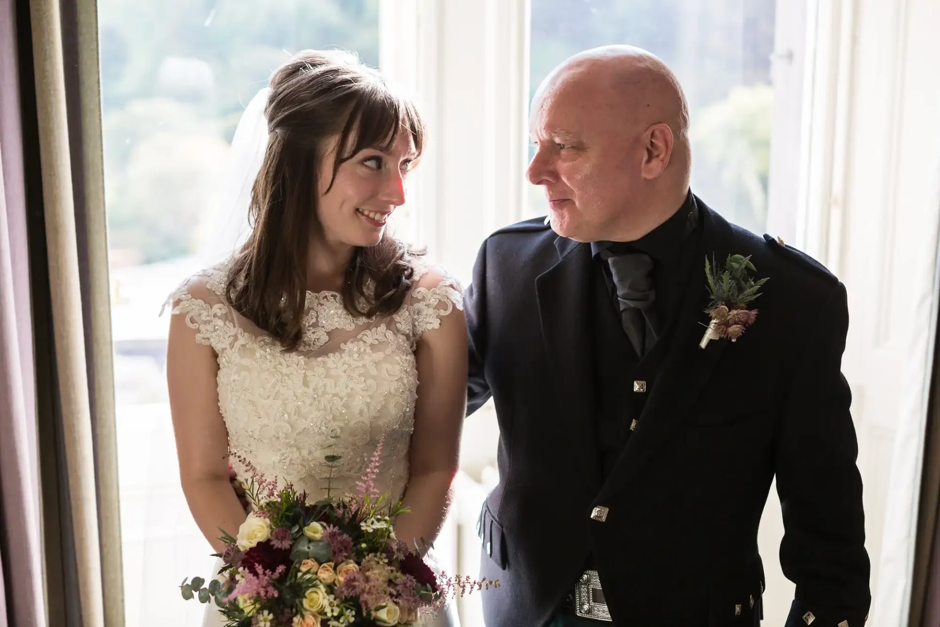 A bride in a lace dress and a man in a black suit with a boutonniere share a joyful glance indoors by a window.