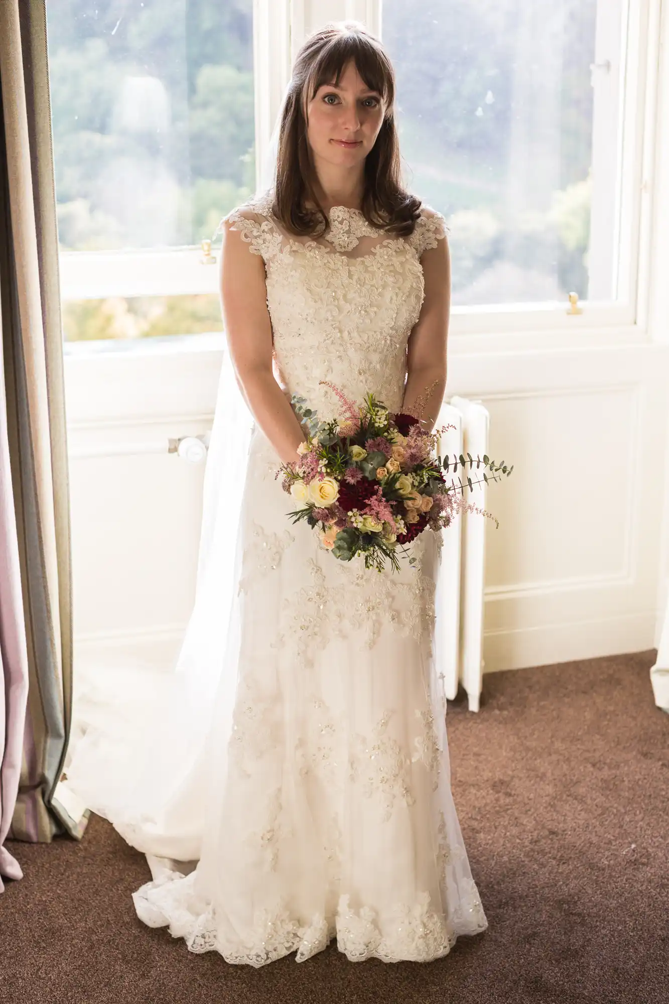 A woman in a white lace wedding dress stands holding a bouquet, with a window behind her casting natural light.