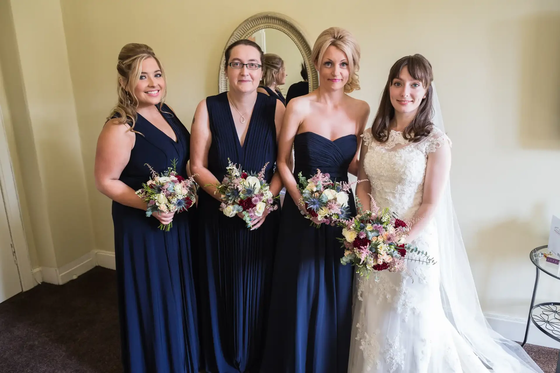 Four women in formal attire, three in navy dresses and one in a bridal gown, holding bouquets and standing in a room with a mirror behind them.