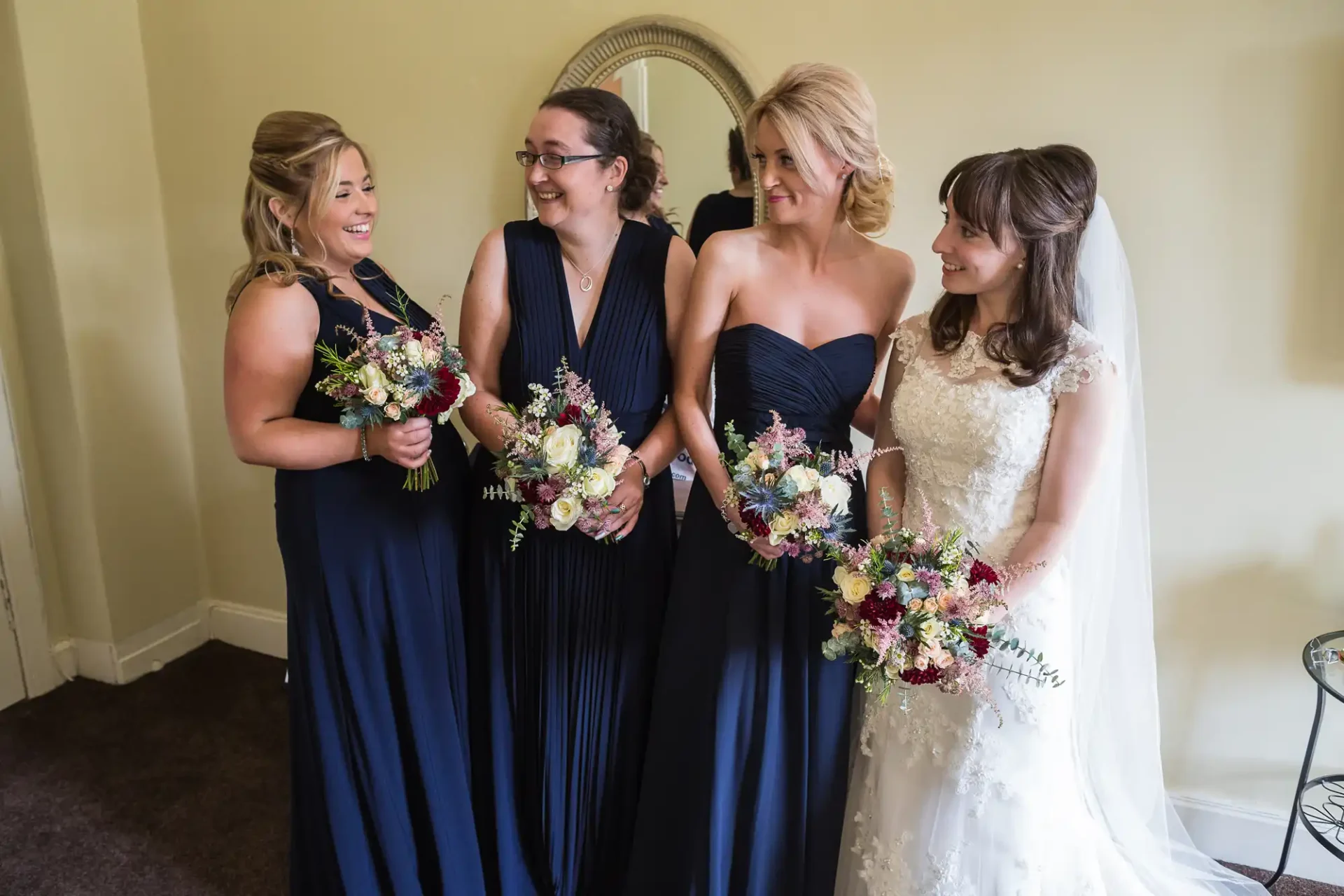 Four women in formal wear, one in a white wedding dress and three in navy dresses, holding bouquets and smiling indoors.