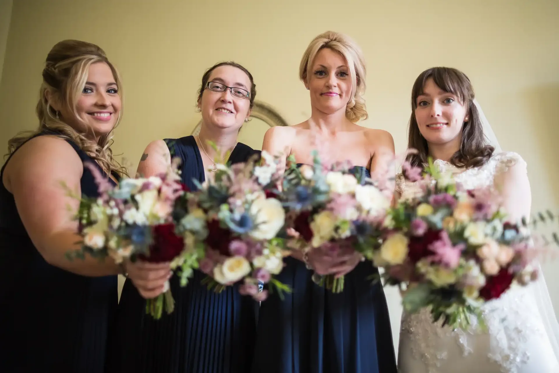 Four women in formal attire, one in a white bridal dress and others in dark dresses, smiling and holding floral bouquets.
