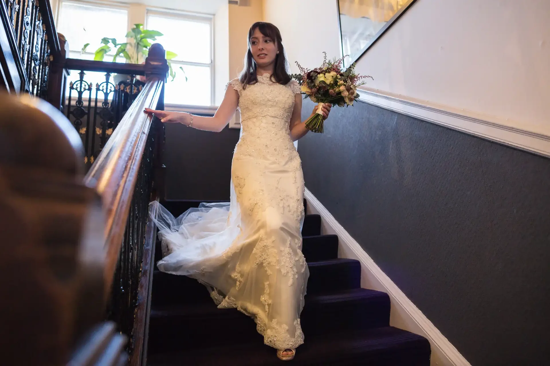 A bride in a white lace dress descends a staircase holding a bouquet, looking towards the camera with a slight smile.