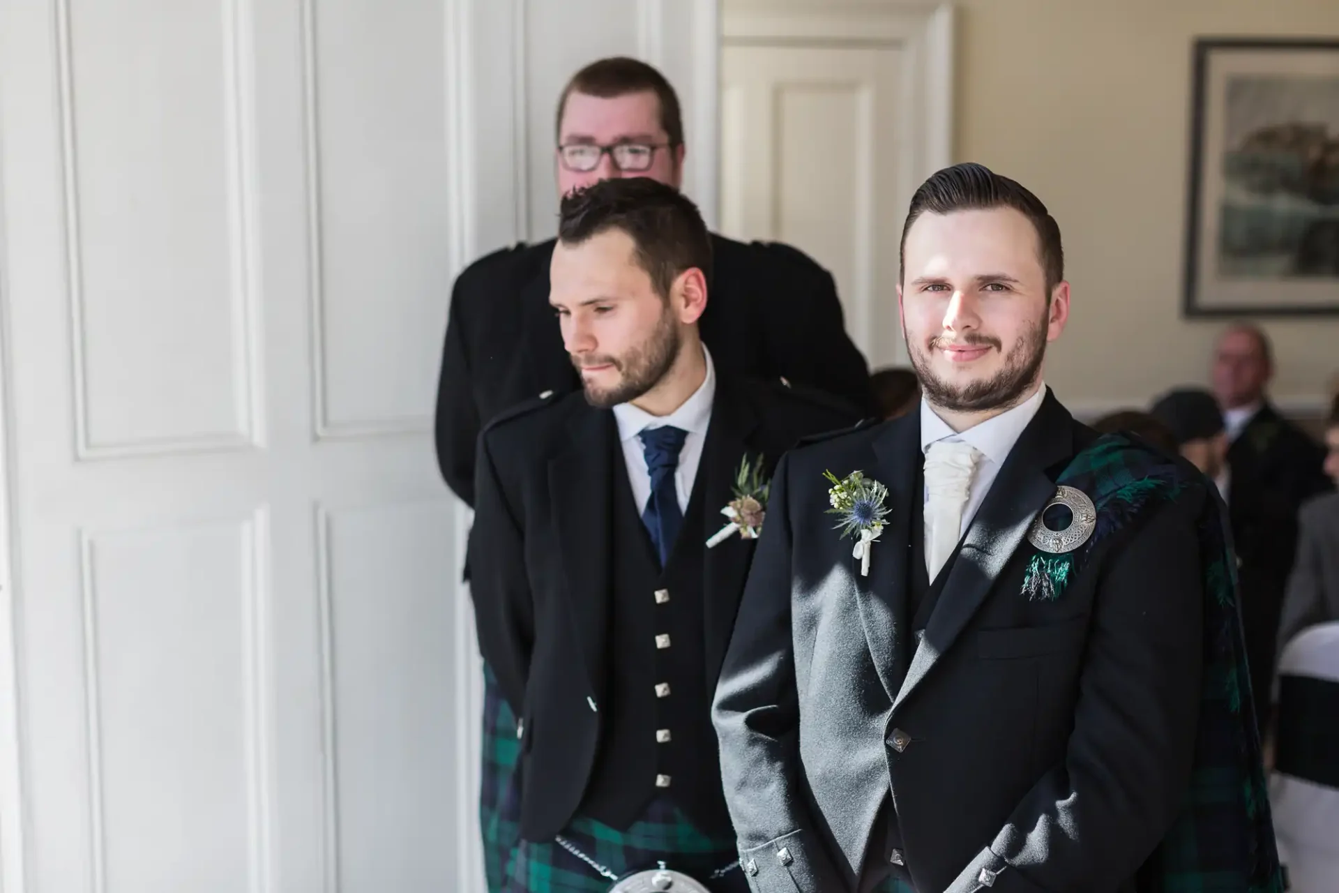 Two men wearing formal attire with tartan elements at an event, one in a kilt, standing in the forefront, with another man partially visible in the background.