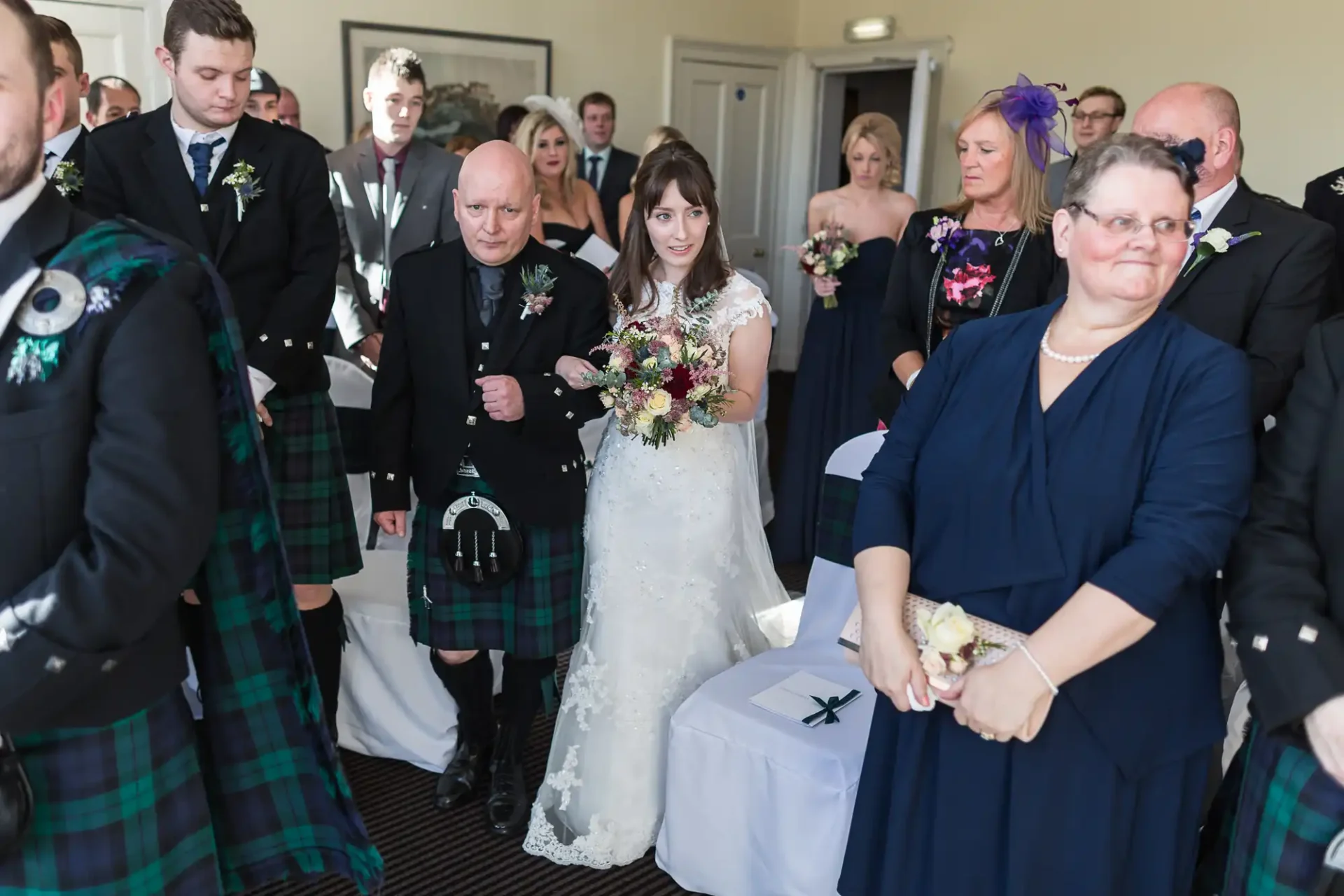 A bride walks down the aisle holding a bouquet, accompanied by her father in a tartan kilt, surrounded by guests in formal attire, at a wedding ceremony.