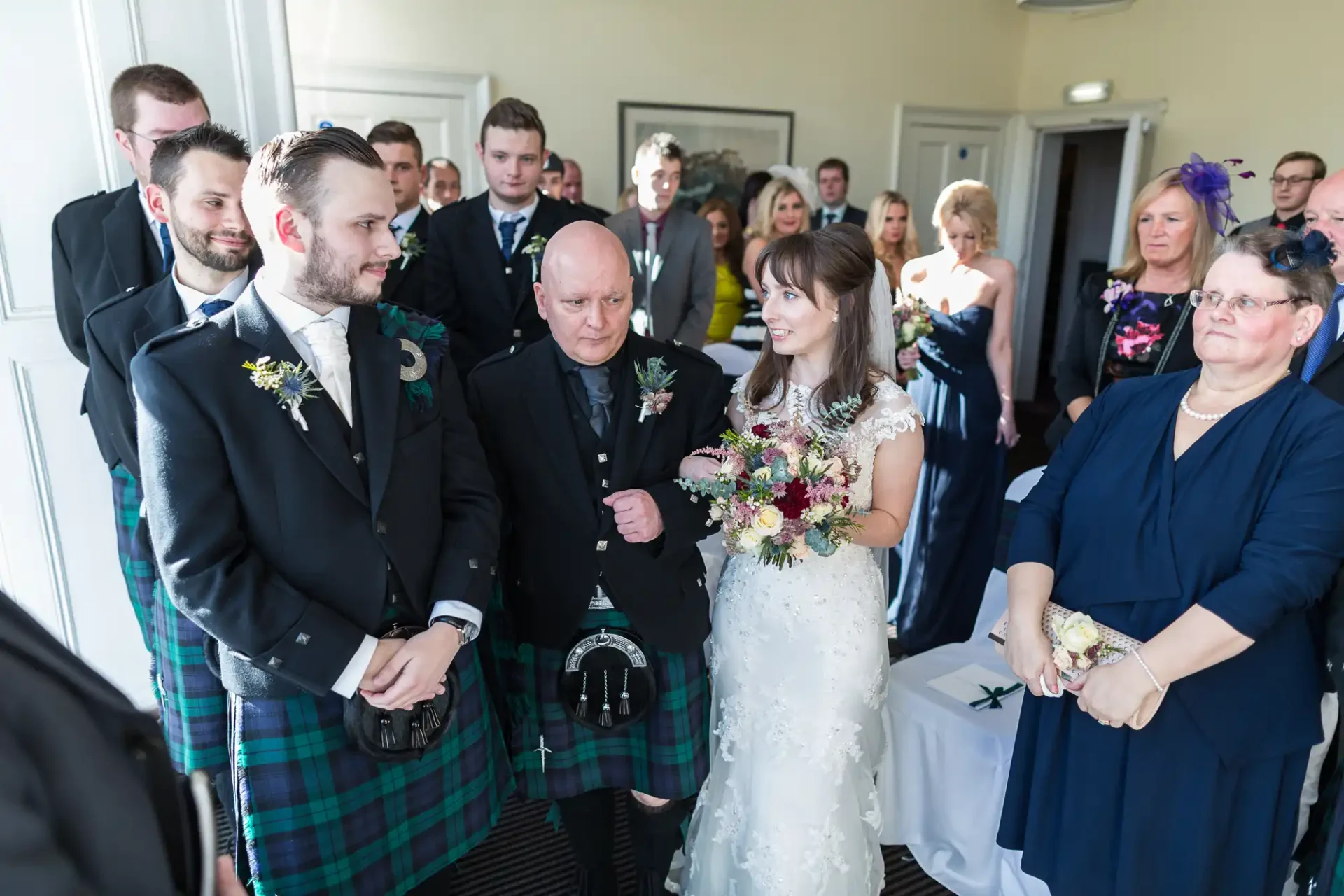 A wedding ceremony with a bride and groom in a kilt surrounded by guests in formal attire, conveying a joyful atmosphere indoors.