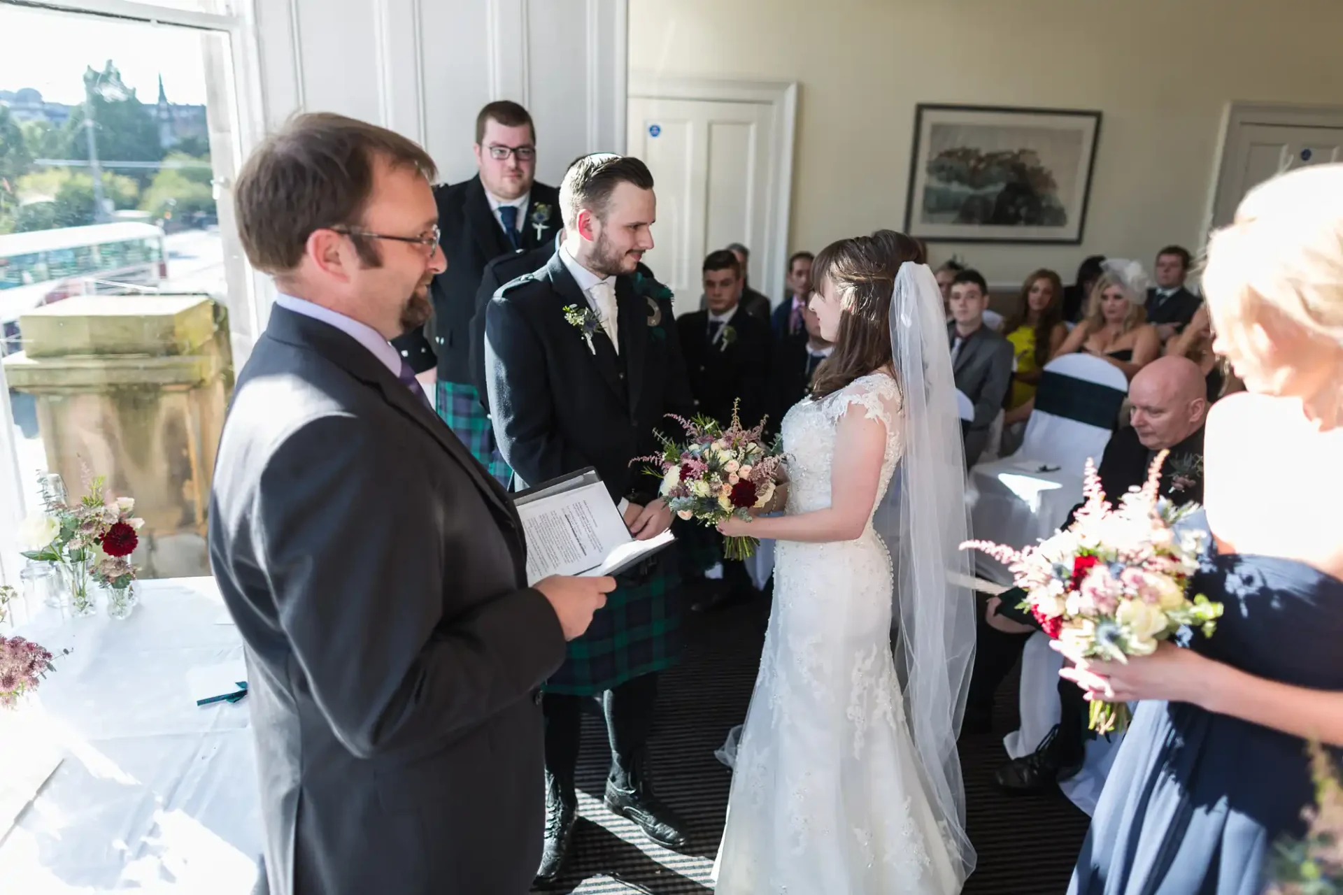 Bride and groom holding hands during a wedding ceremony in a sunlit room, with guests and an officiant surrounding them.