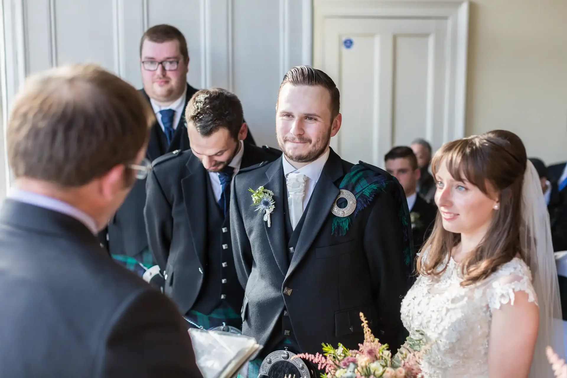 A groom smiling at a speaker during a wedding ceremony, with a bride and groomsmen in background, all dressed formally.