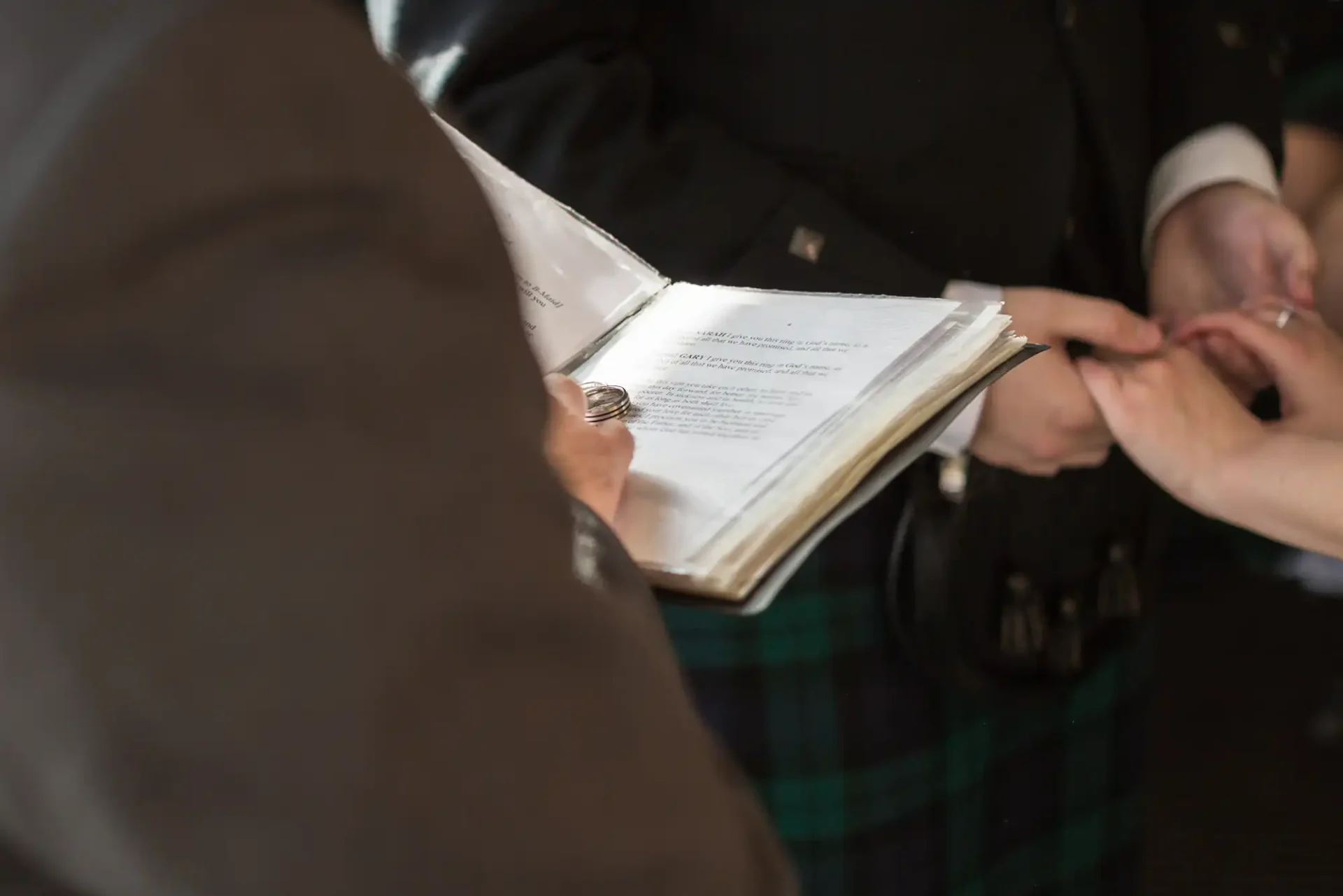 Close-up of a wedding ceremony with hands exchanging rings, officiant holding a book with text, and groom in kilt visible.