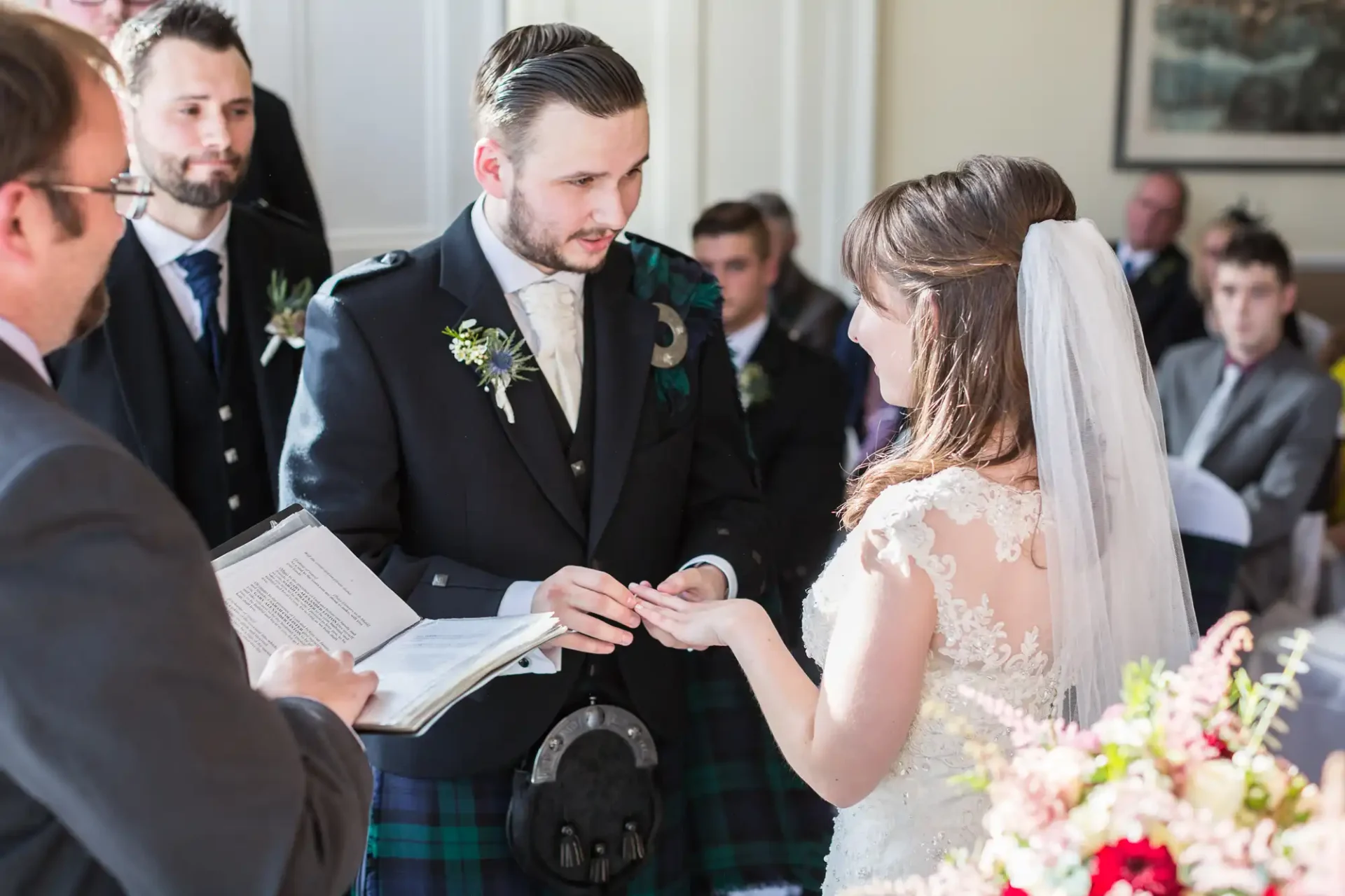 Bride and groom exchanging rings during a wedding ceremony, with the groom wearing a kilt. guests observe the moment.