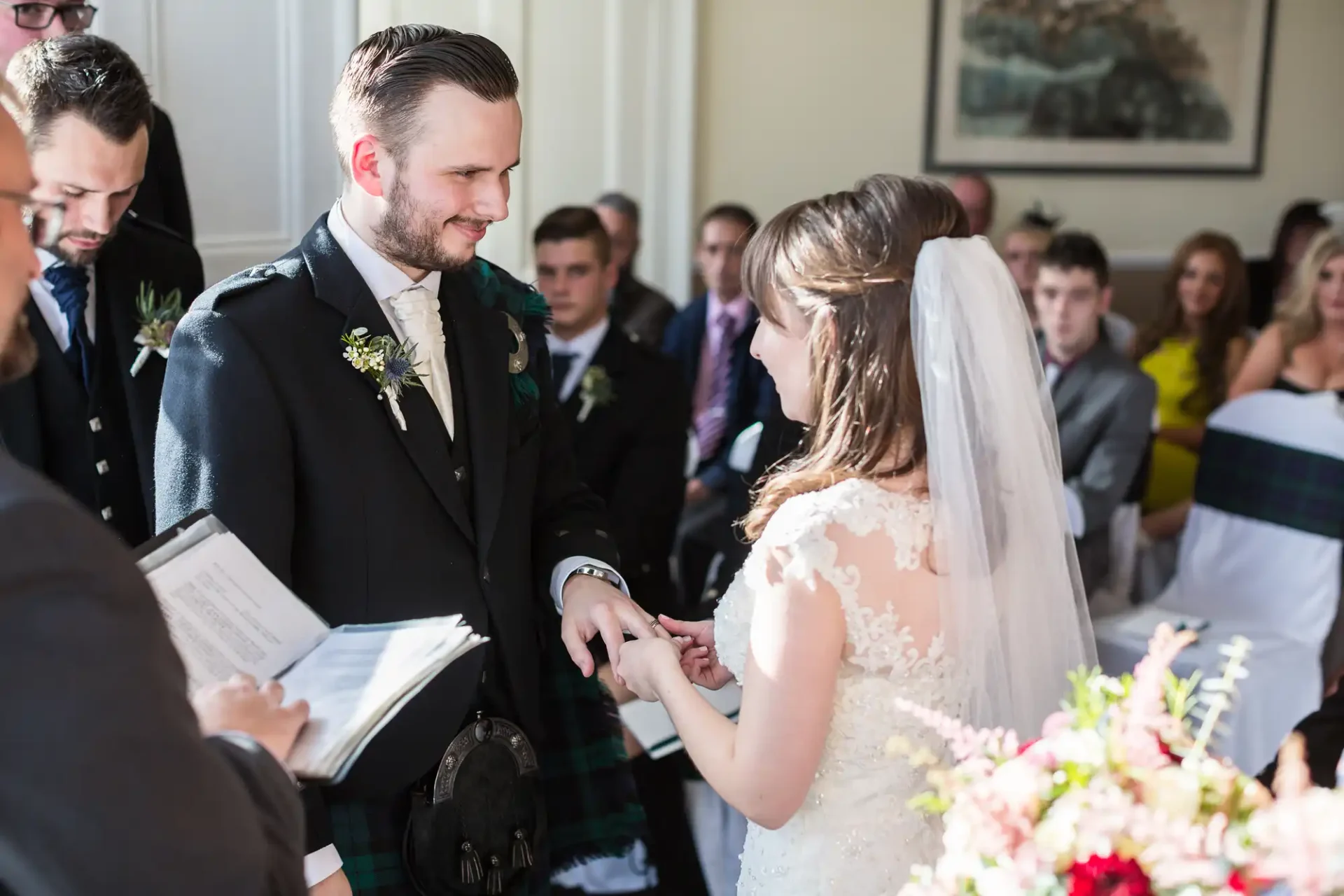 Bride and groom exchanging rings during a wedding ceremony in a room filled with guests, the groom wearing a kilt.