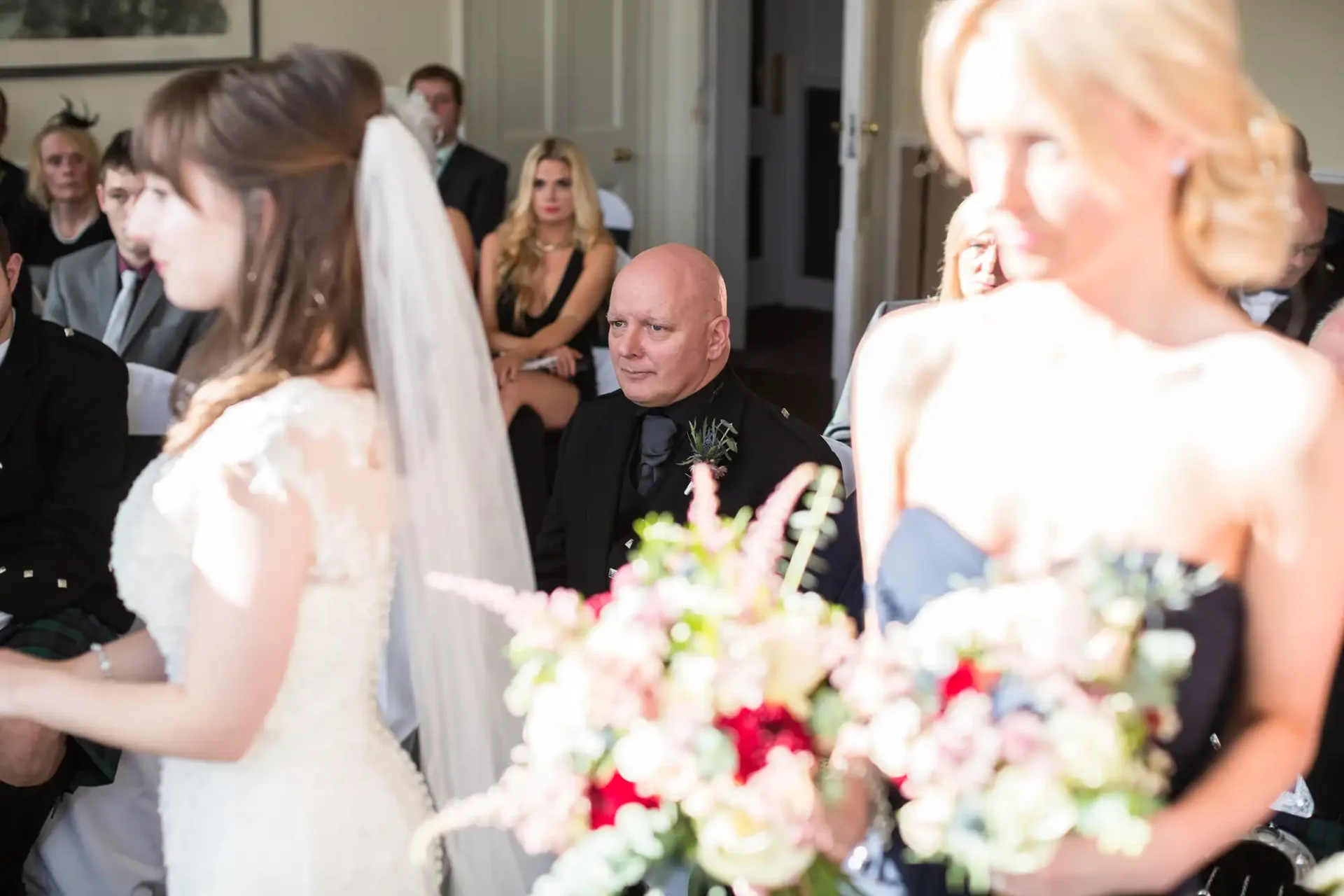 A bride in a white dress and veil walks past seated guests, focusing on a man in the foreground looking reflective.
