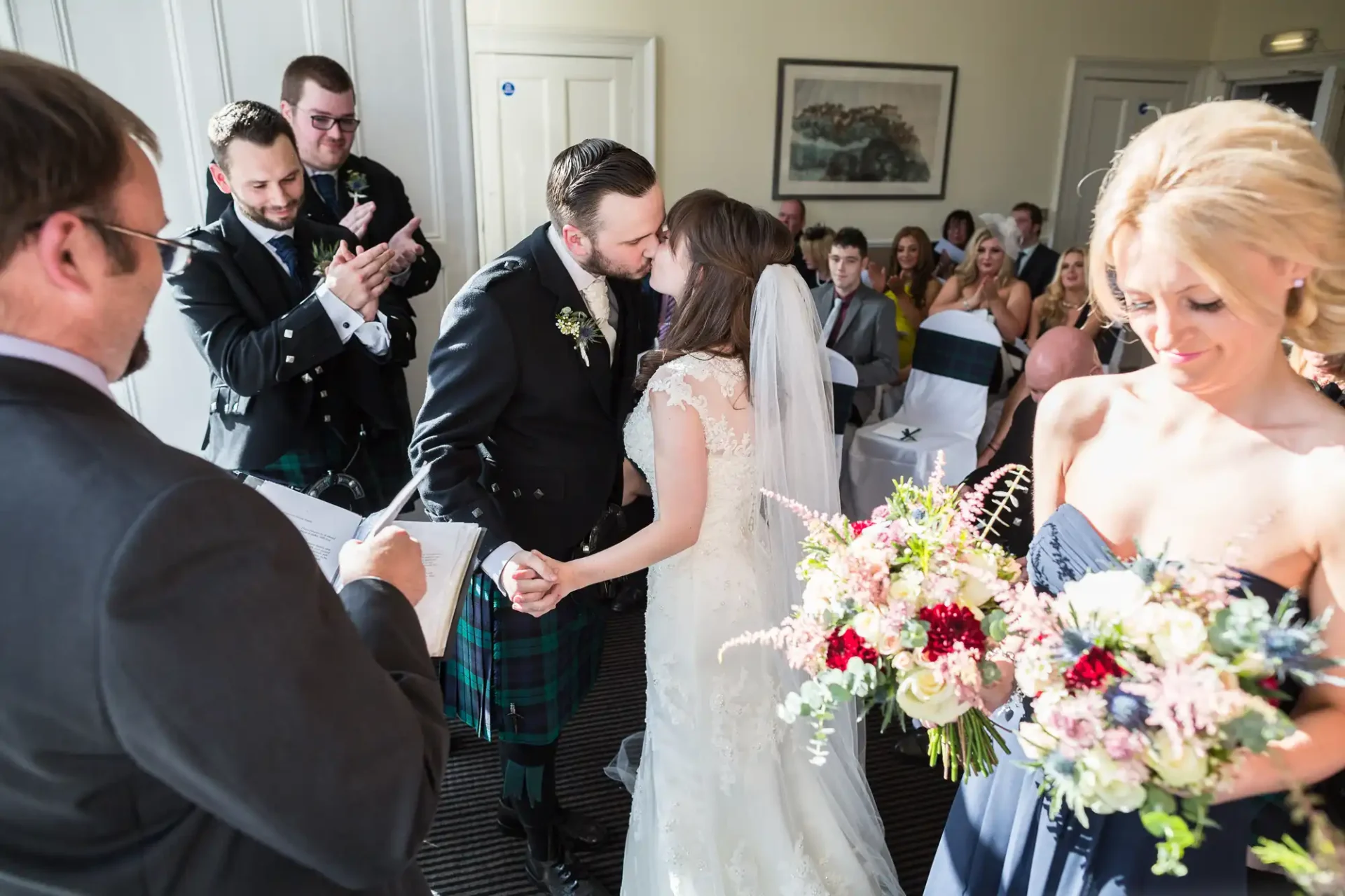 Bride and groom kissing at the altar, surrounded by guests, with a man in a kilt and a woman holding a floral bouquet visible.
