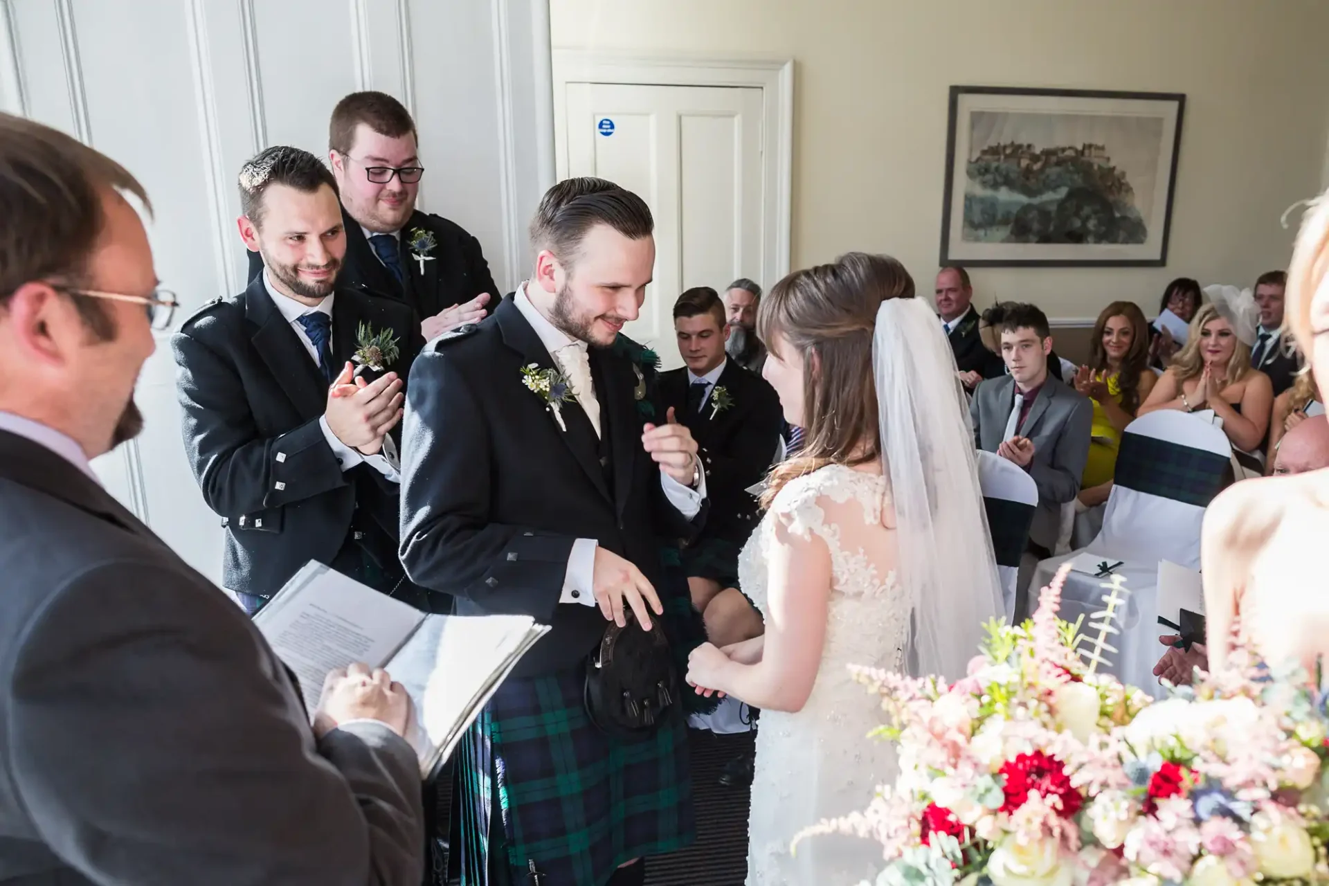 A bride and groom exchanging wedding rings in a crowded room, groom wearing a kilt, with smiling guests looking on.