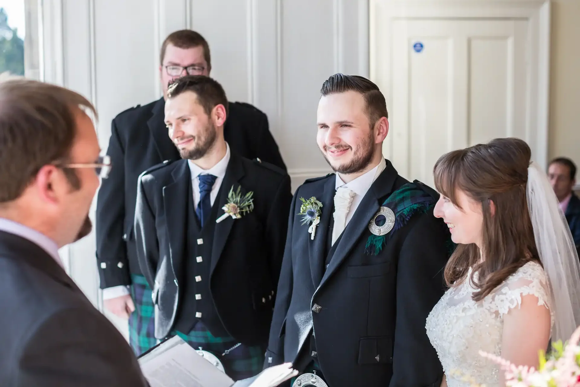 A wedding ceremony with a smiling groom in a kilt and bride in a white dress, standing with guests in a well-lit room.
