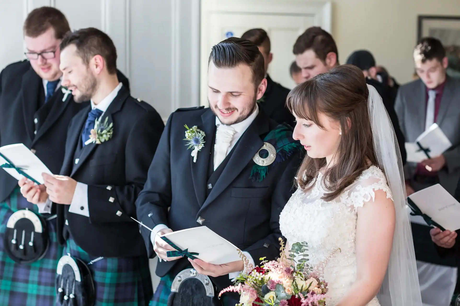 A bride and groom in wedding attire reading from a booklet alongside groomsmen in kilts and suits.