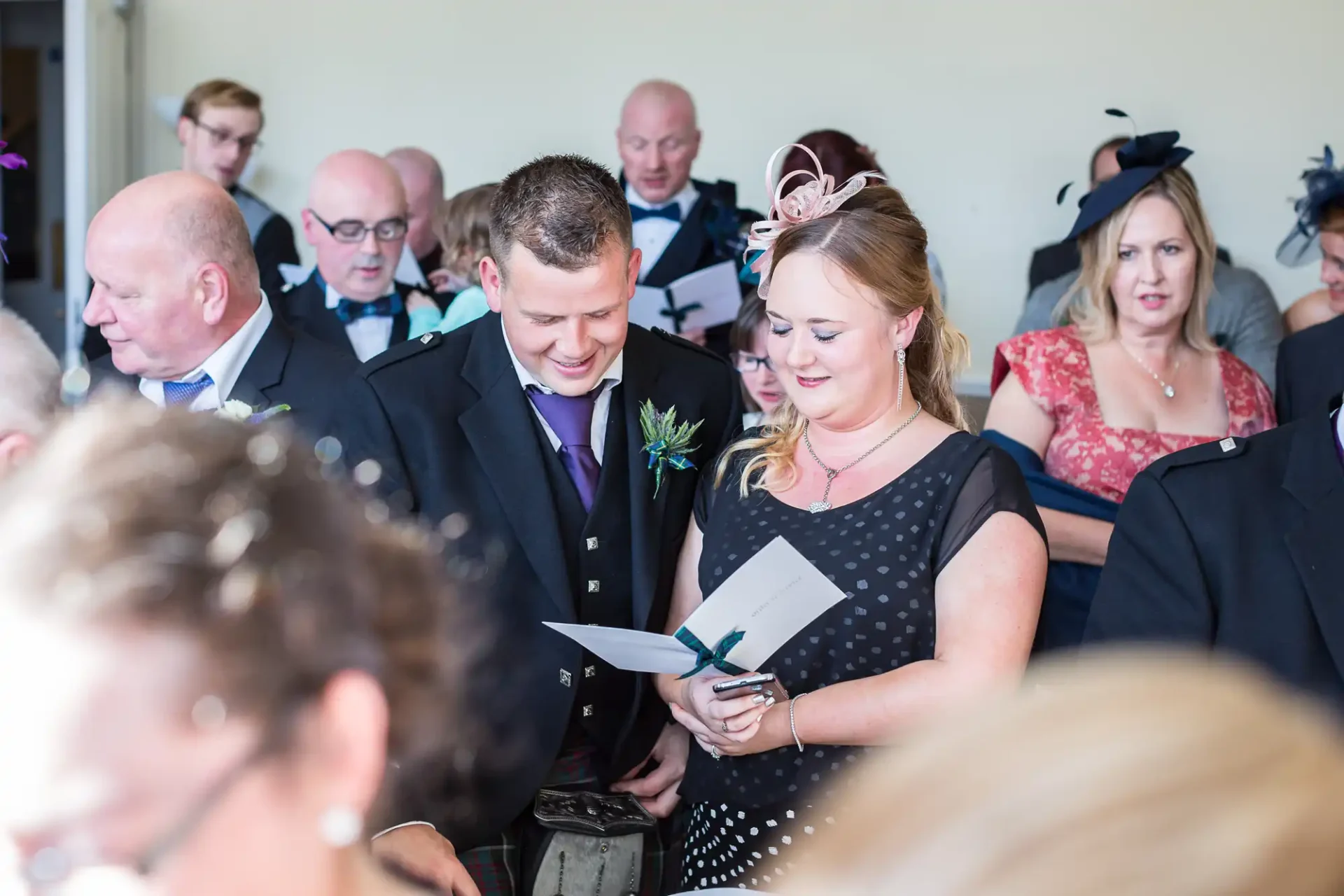 A couple reads a document together at a wedding, surrounded by guests in formal attire.