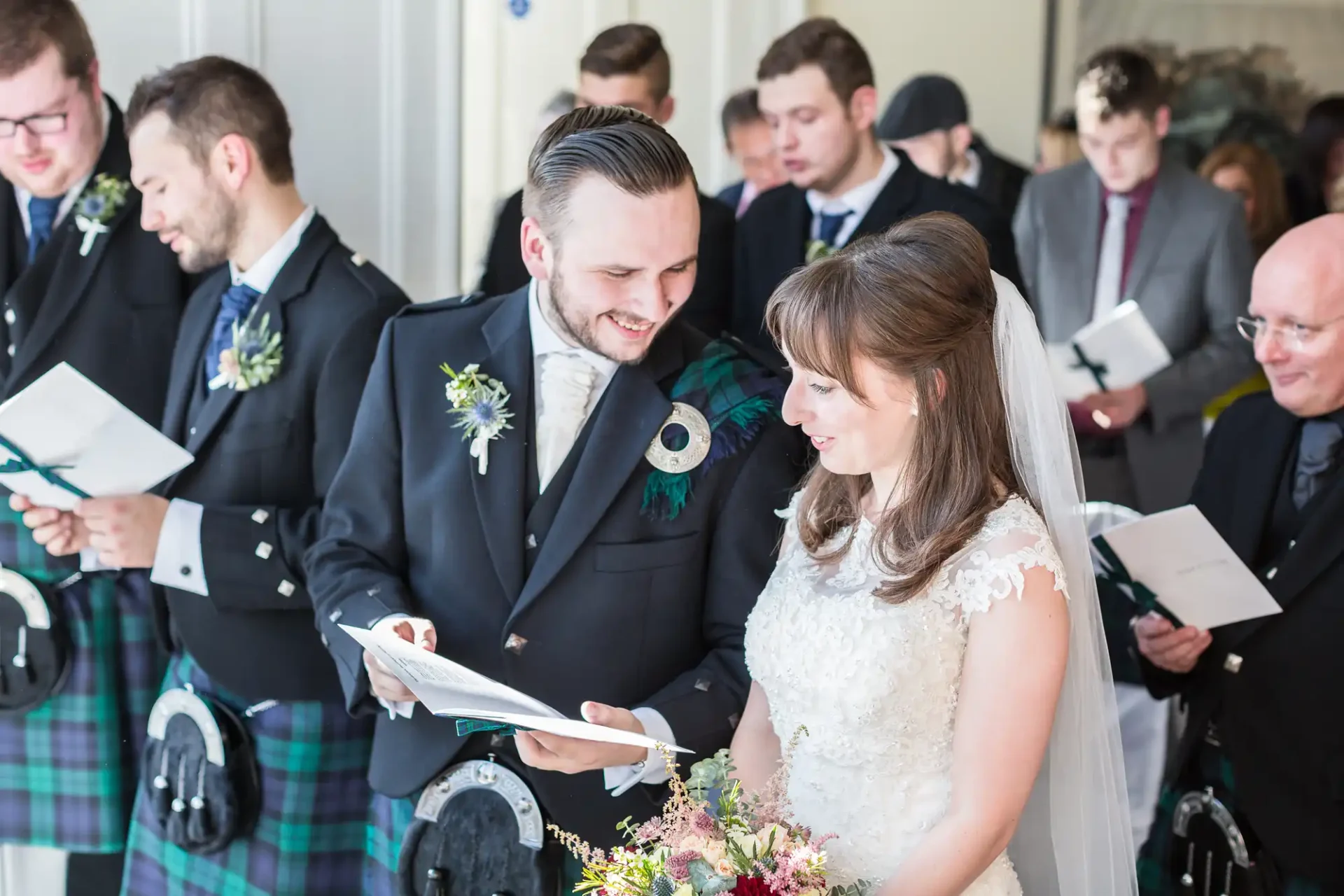Bride and groom smiling and reading from a paper in a wedding ceremony, surrounded by guests in formal attire.