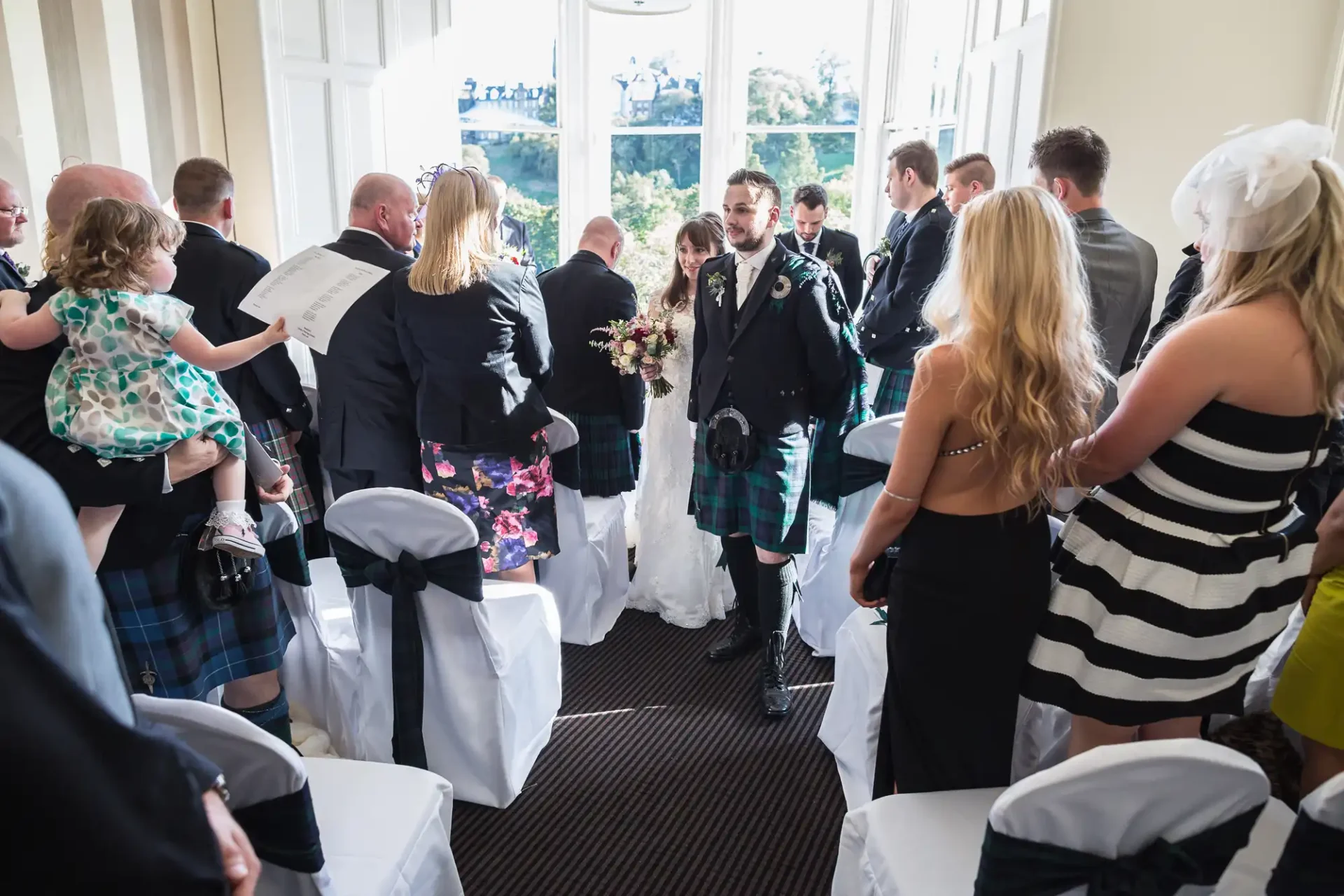 A wedding reception in a bright room with guests in formal attire, some wearing kilts; a couple walks down the aisle, greeted warmly by attendees.