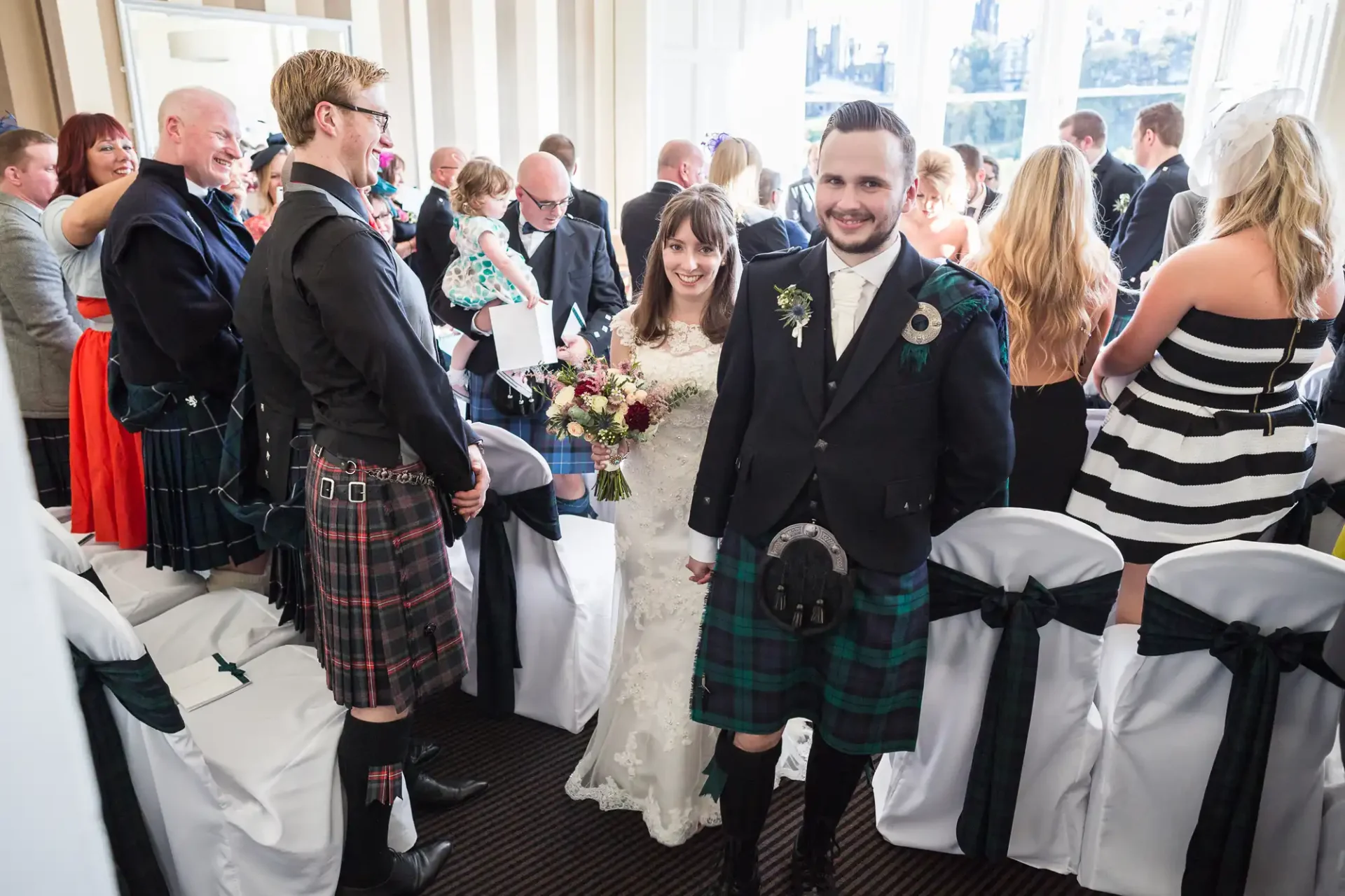 A bride and groom, both in traditional scottish attire, smiling as they walk through a crowded room of wedding guests.