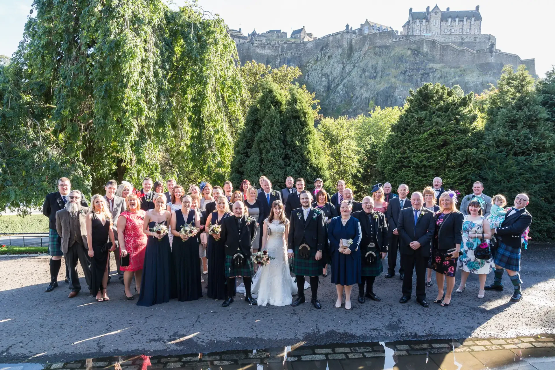 A large group of people in formal attire posing for a photo at a wedding, with a historic castle in the background on a sunny day.